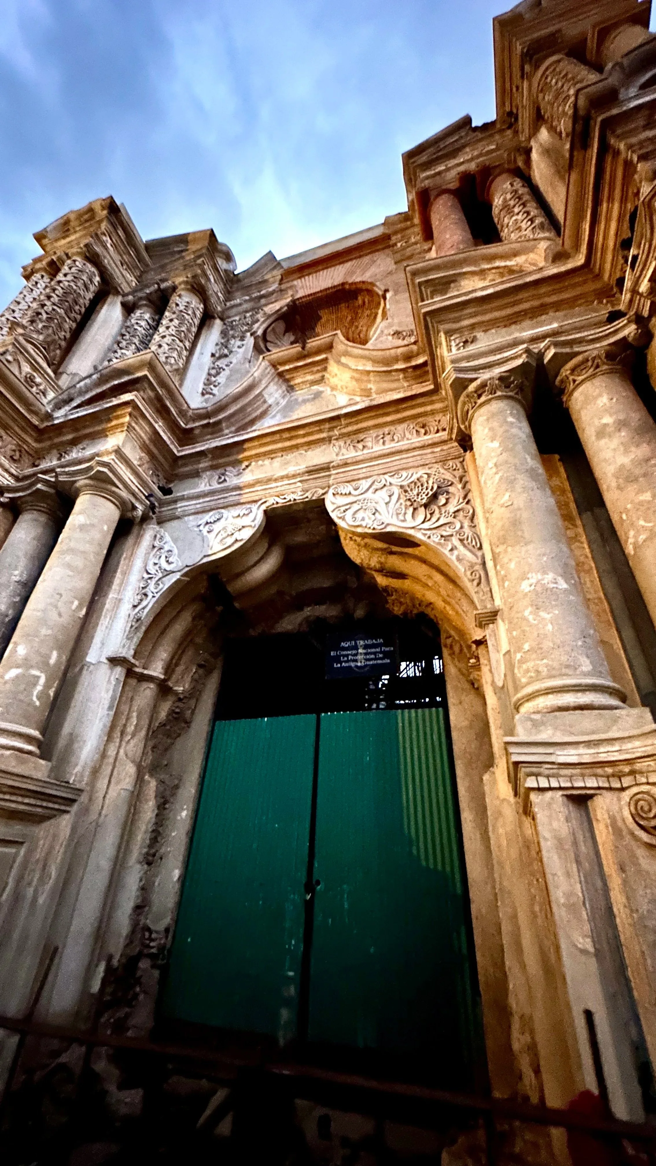 A low-angle view of an antique stone building with ornate architectural details and tall columns features a weathered green door at the entrance. in Antigua, Guatemala. 