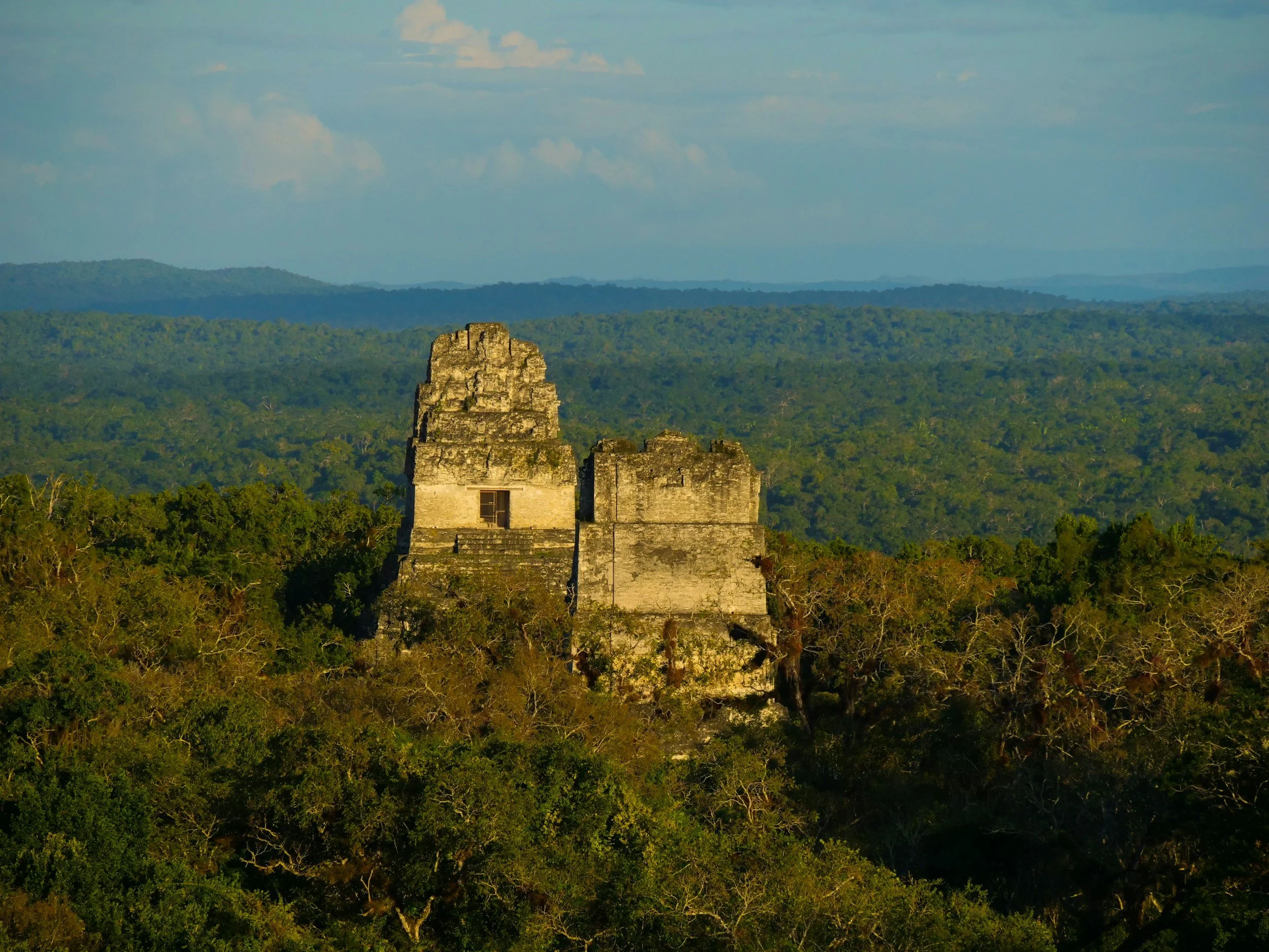 An ancient stone structure, possibly a Mayan temple, surrounded by lush green forest with hills in the distance and a partly cloudy sky.