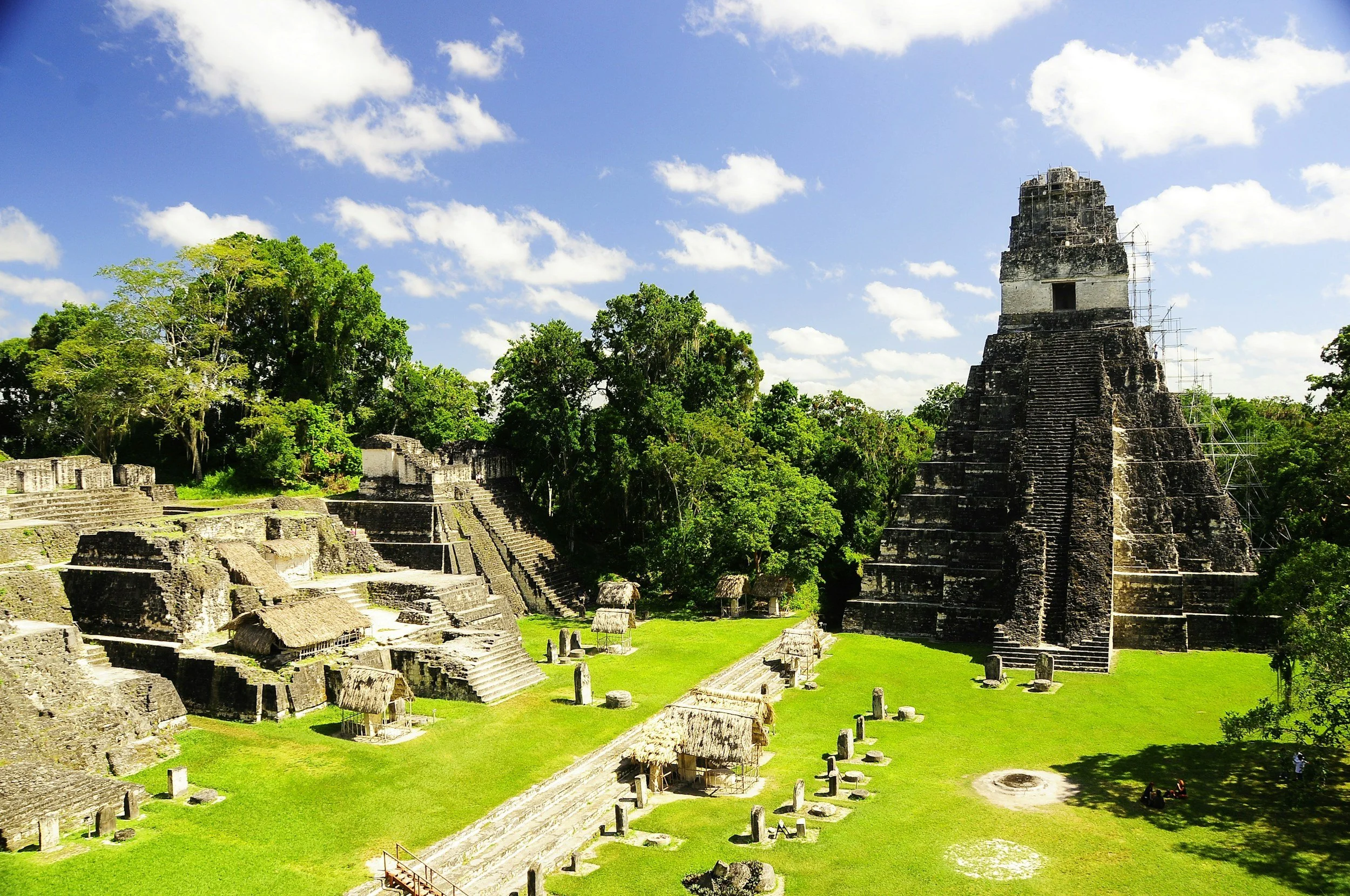 Ancient Mayan ruins with a stepped pyramid and other stone structures, surrounded by lush green trees under a blue sky with scattered clouds.