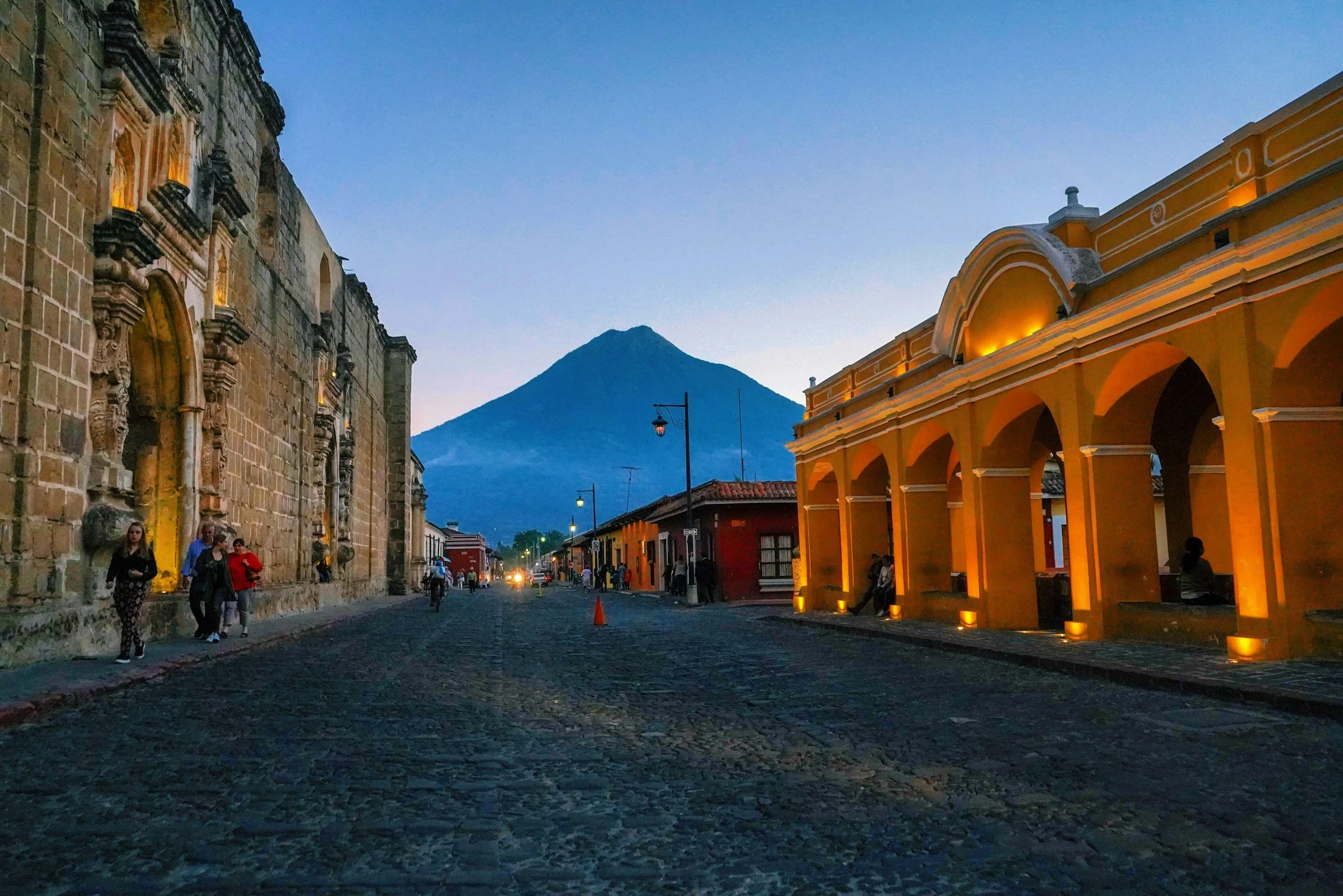City street scene at dusk with historic buildings on either side, a mountain in the background, and people walking along the cobblestone street.