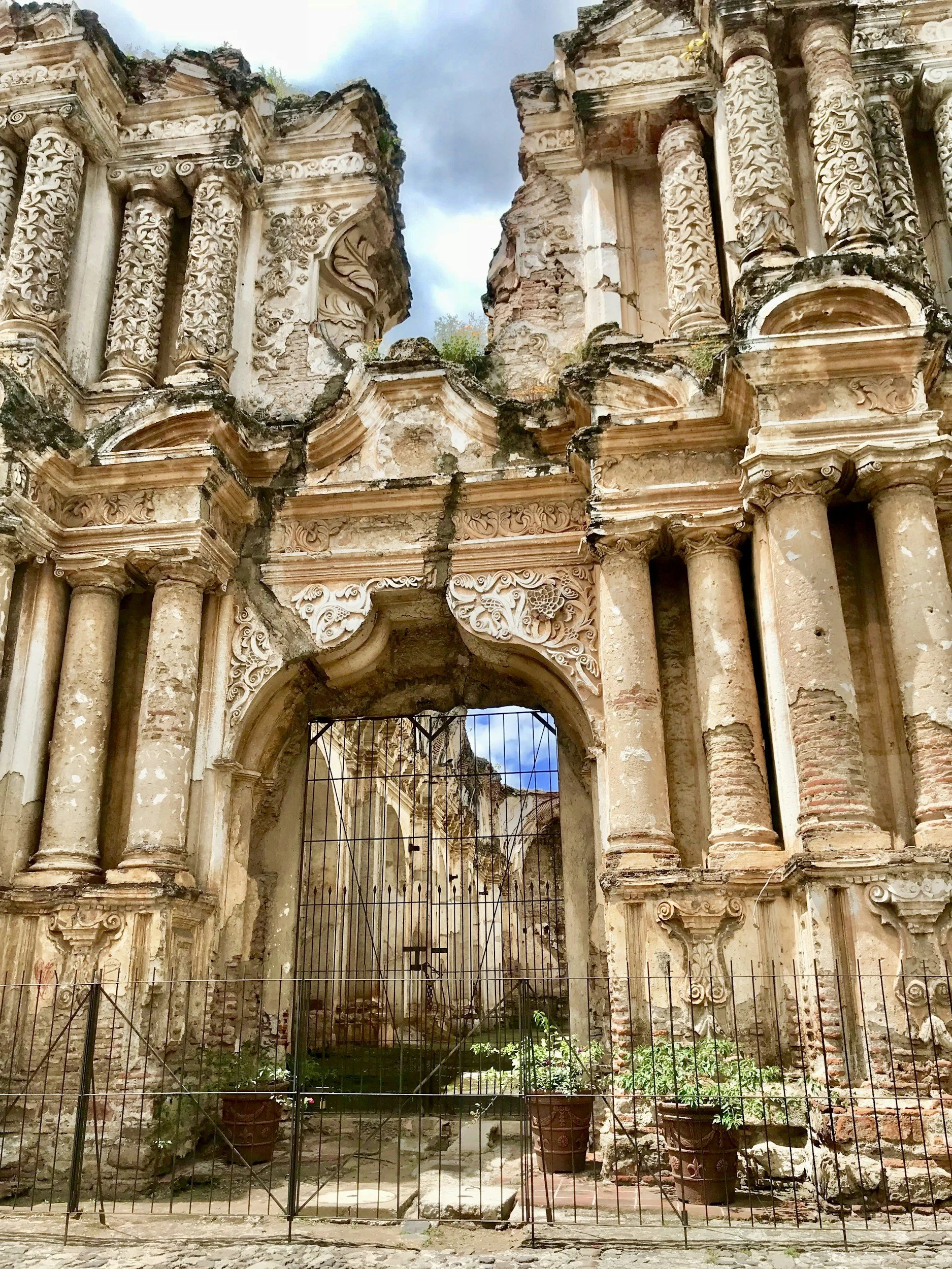 Ruins of an ancient building with ornate carvings and columns, partly collapsed, with a metal gate in front and potted plants at the base.