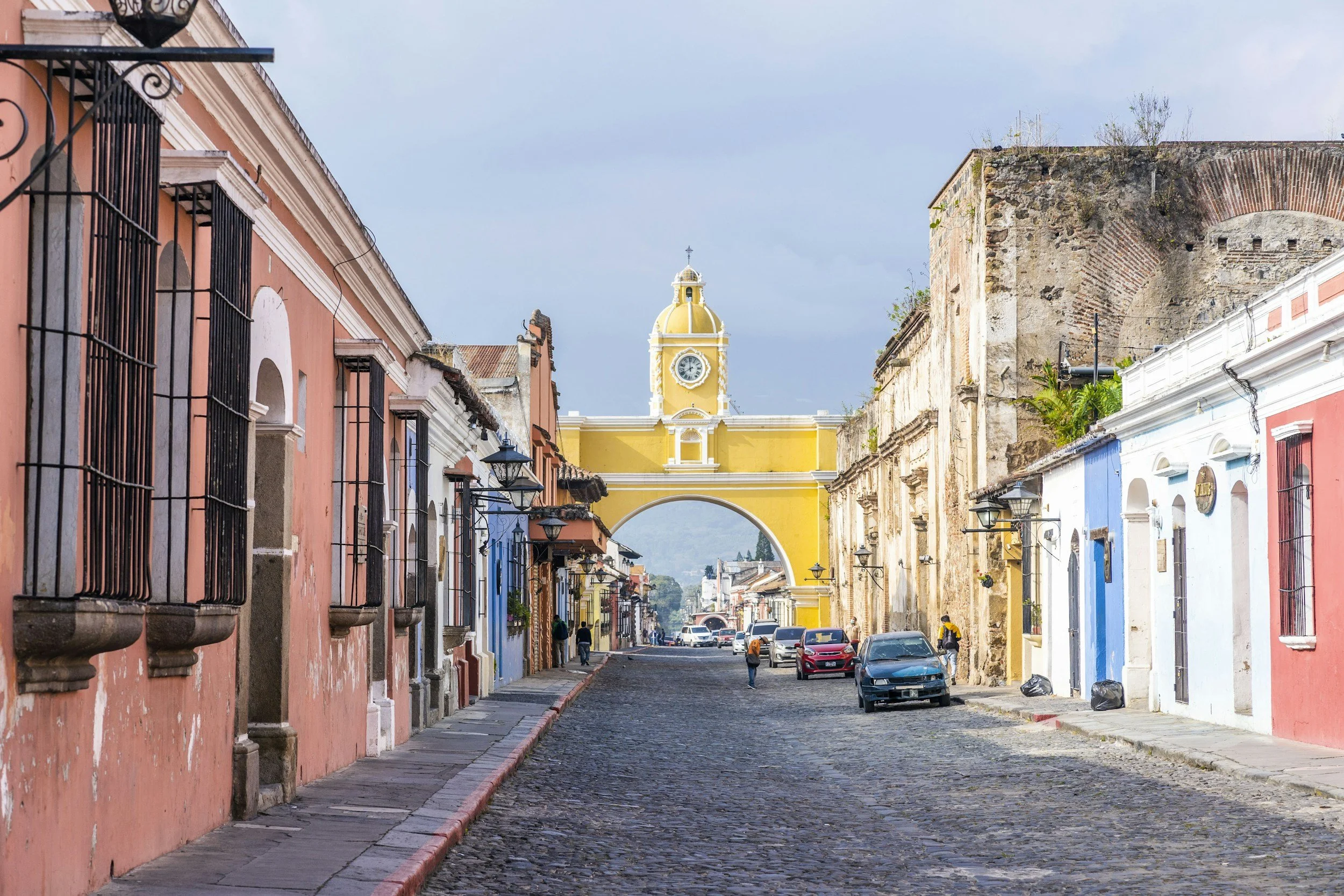 Colorful colonial-style buildings line a cobblestone street, leading to a yellow church with a clock tower and a domed roof in the background under a partly cloudy sky. People walk along the street, and cars are parked nearby.