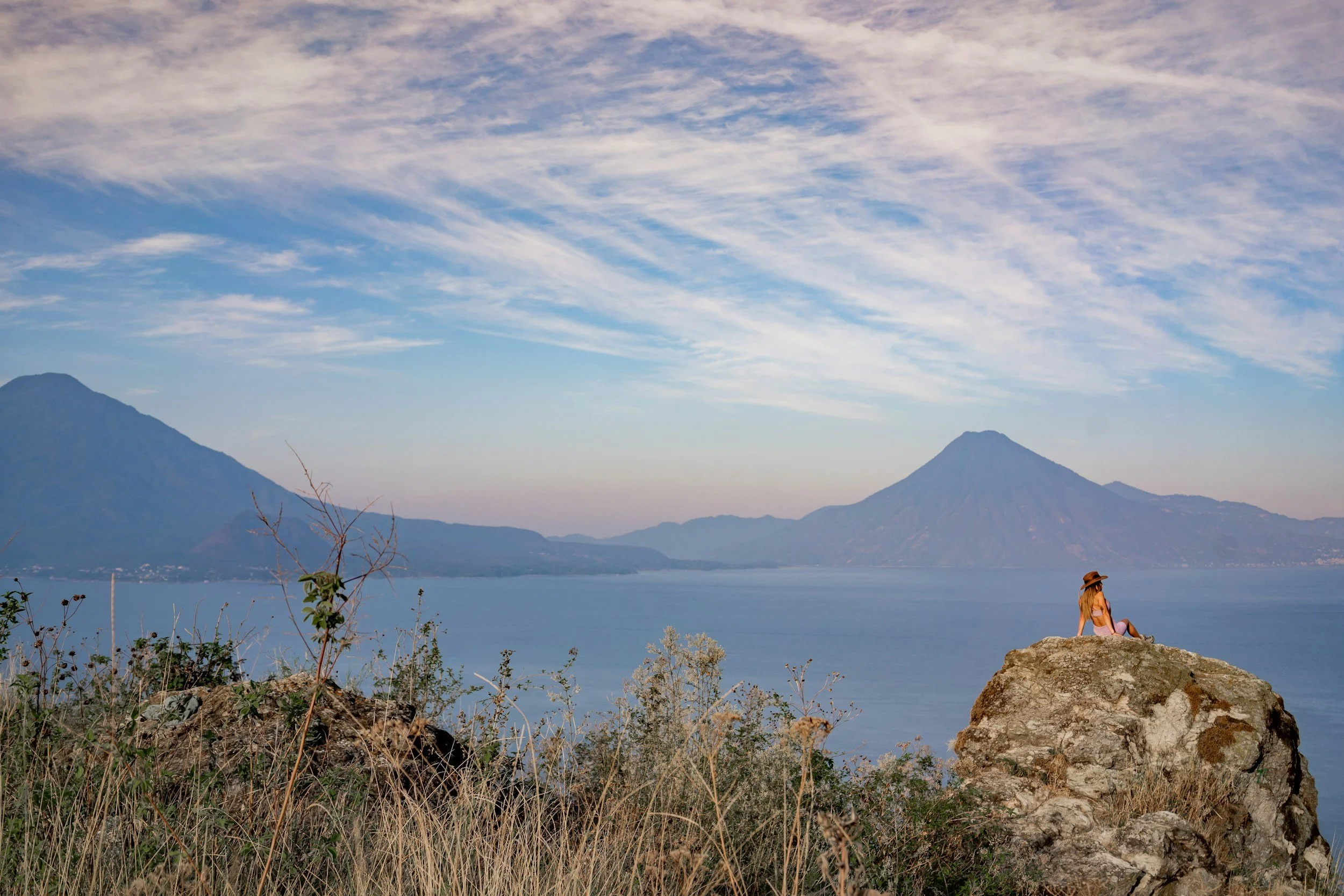 A woman sitting on a large rock by Lake Atitlan in Guatemala with volcano views in the background, wearing a hat and swimsuit, under a partly cloudy sky.