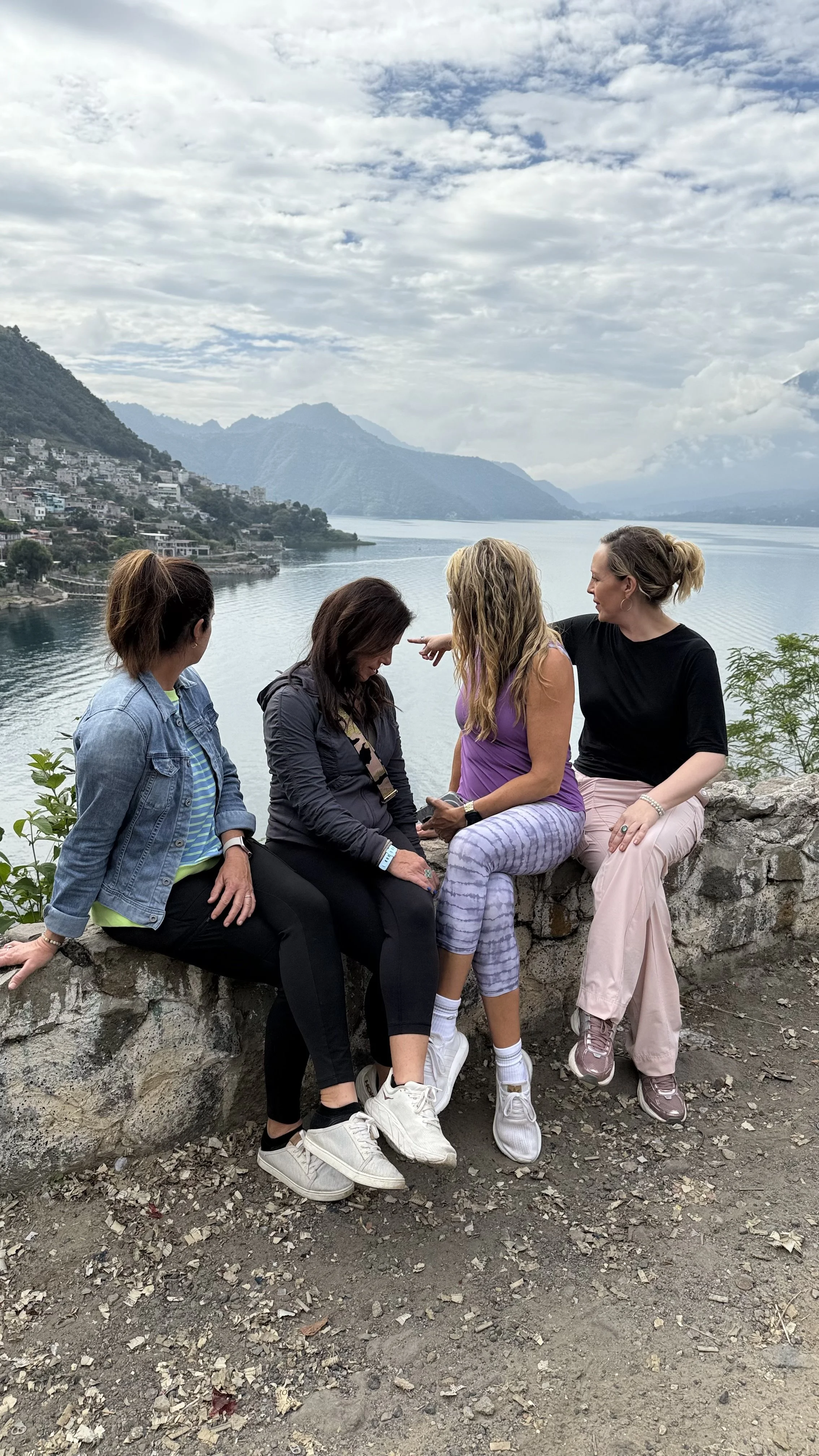 Four women sitting on a stone wall overlooking Lake Atitlan in Guatemala with volcanoes in the background, engaged in conversation.