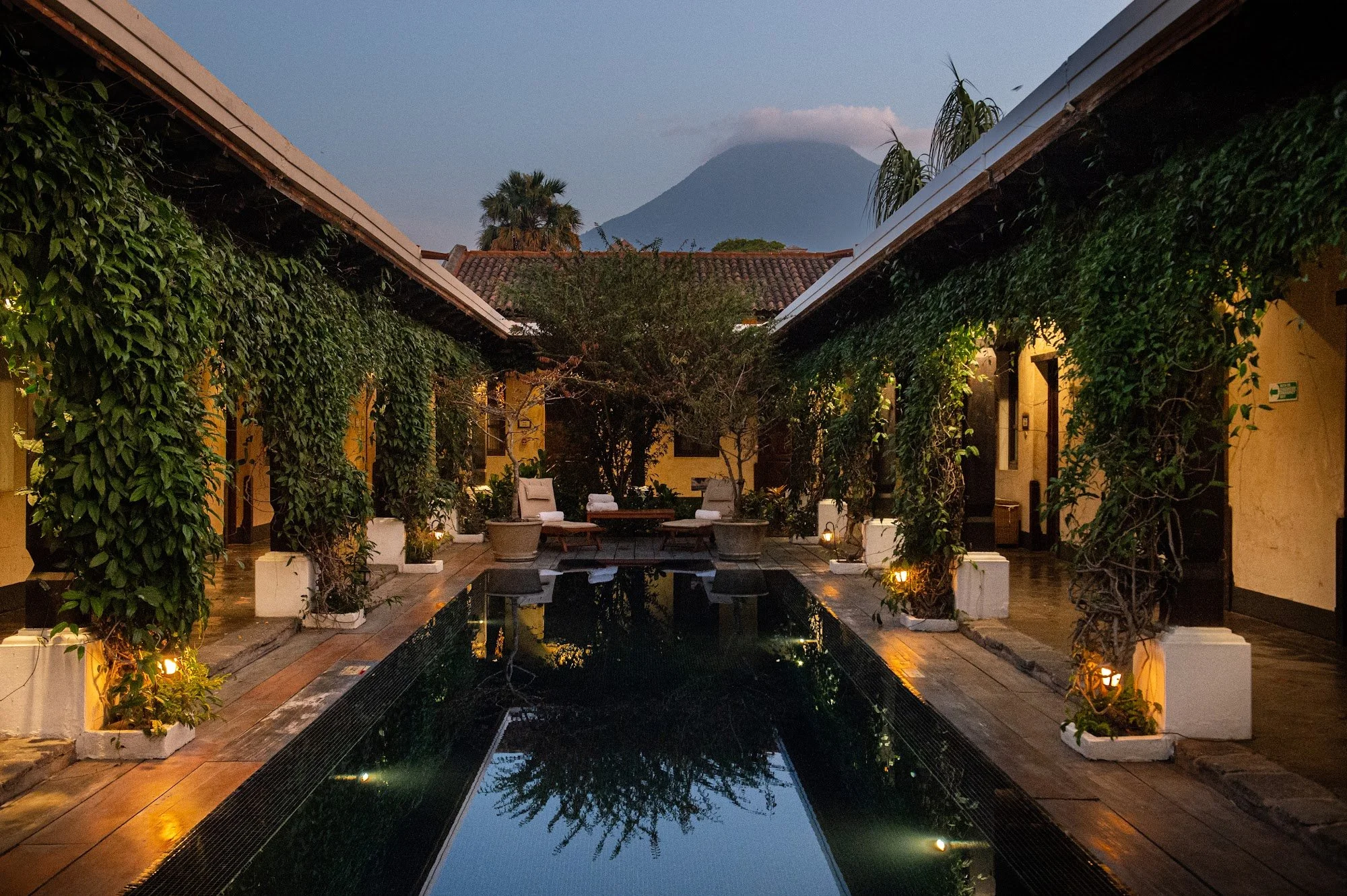 A courtyard with a reflecting pool, surrounded by chairs and lush greenery, with mountain in the background during dusk.
