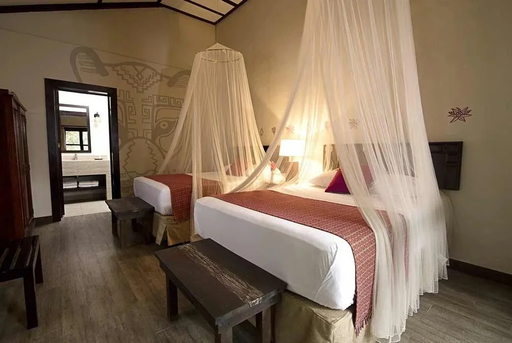 Hotel room with two beds covered by white mosquito netting, a nightstand with a lamp between the beds, a wooden bench at the foot of each bed, a wooden wardrobe, and a doorway leading to a bathroom with a sink and mirror.