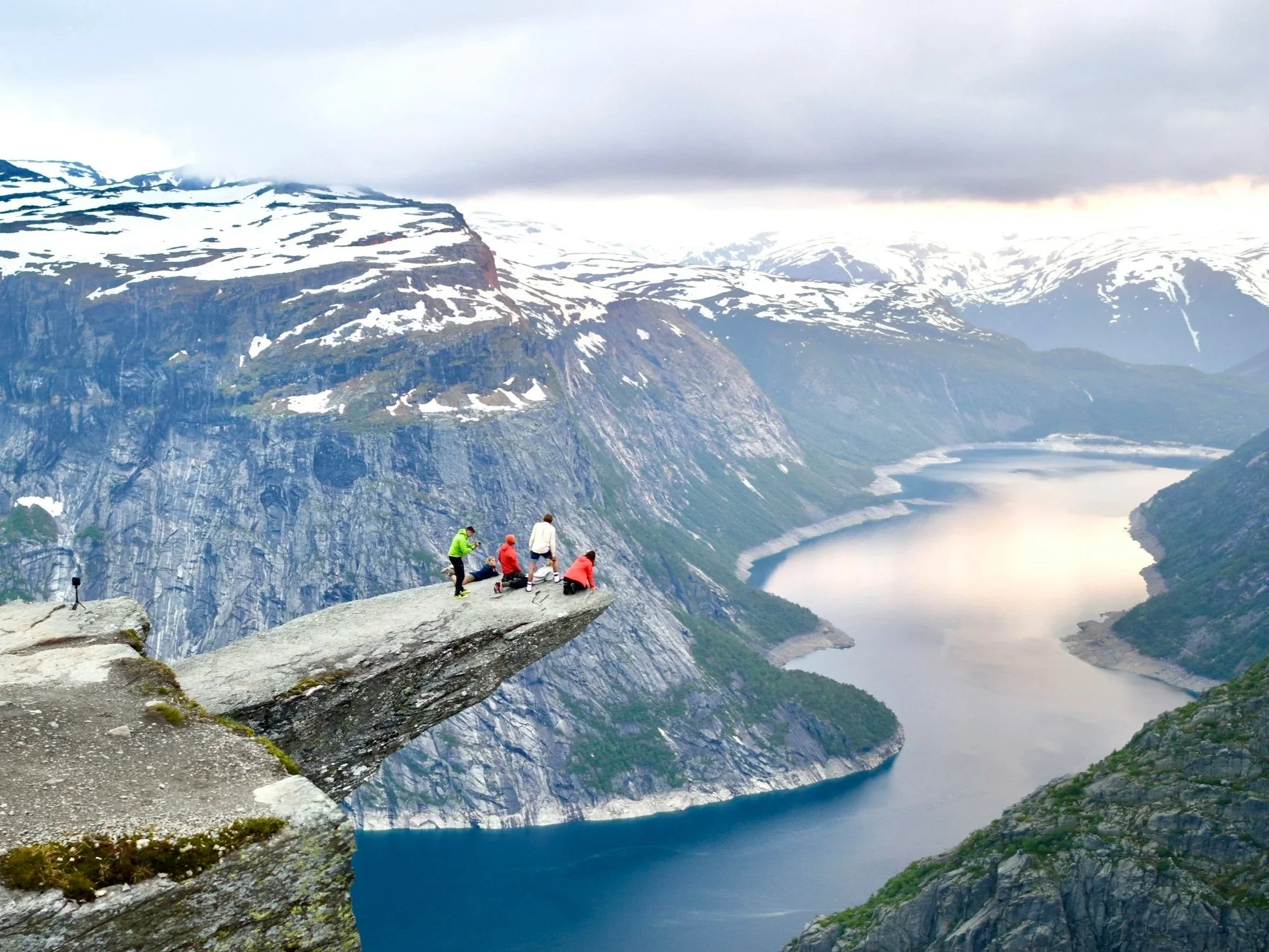 A group of seven hikers on a rocky ledge overlooking a fjord with steep, snow-capped mountains in the background, under a cloudy sky.