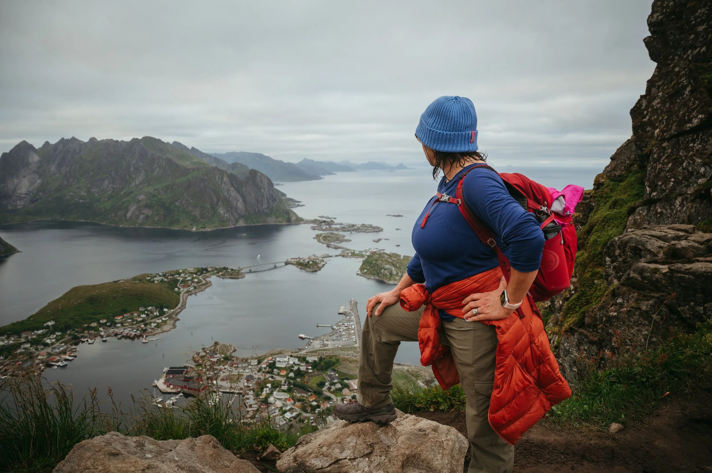 A woman wearing a blue beanie, blue long-sleeve shirt, khaki pants, and hiking boots, with a red backpack and orange jacket tied around her waist, sits on a rock looking out over a coastal town, water, mountains, and cloudy sky.