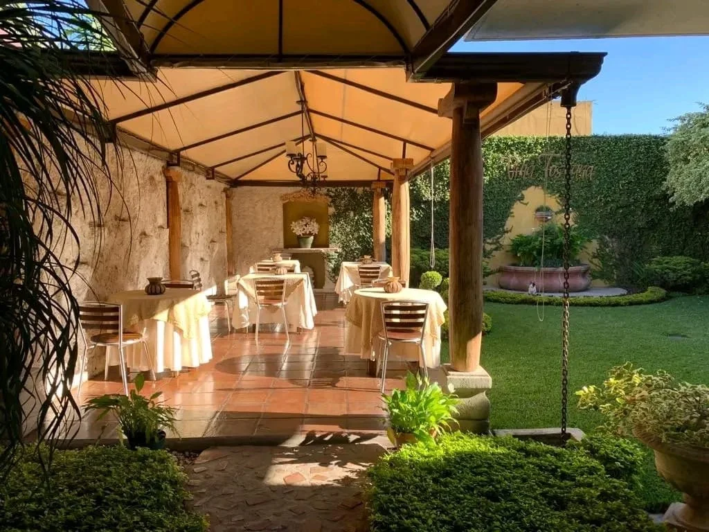 Outdoor dining area with round tables covered in yellow tablecloths, set under a canopy with support beams. There are chairs around each table. The area is surrounded by greenery, including grass, plants, and a fountain in the background.