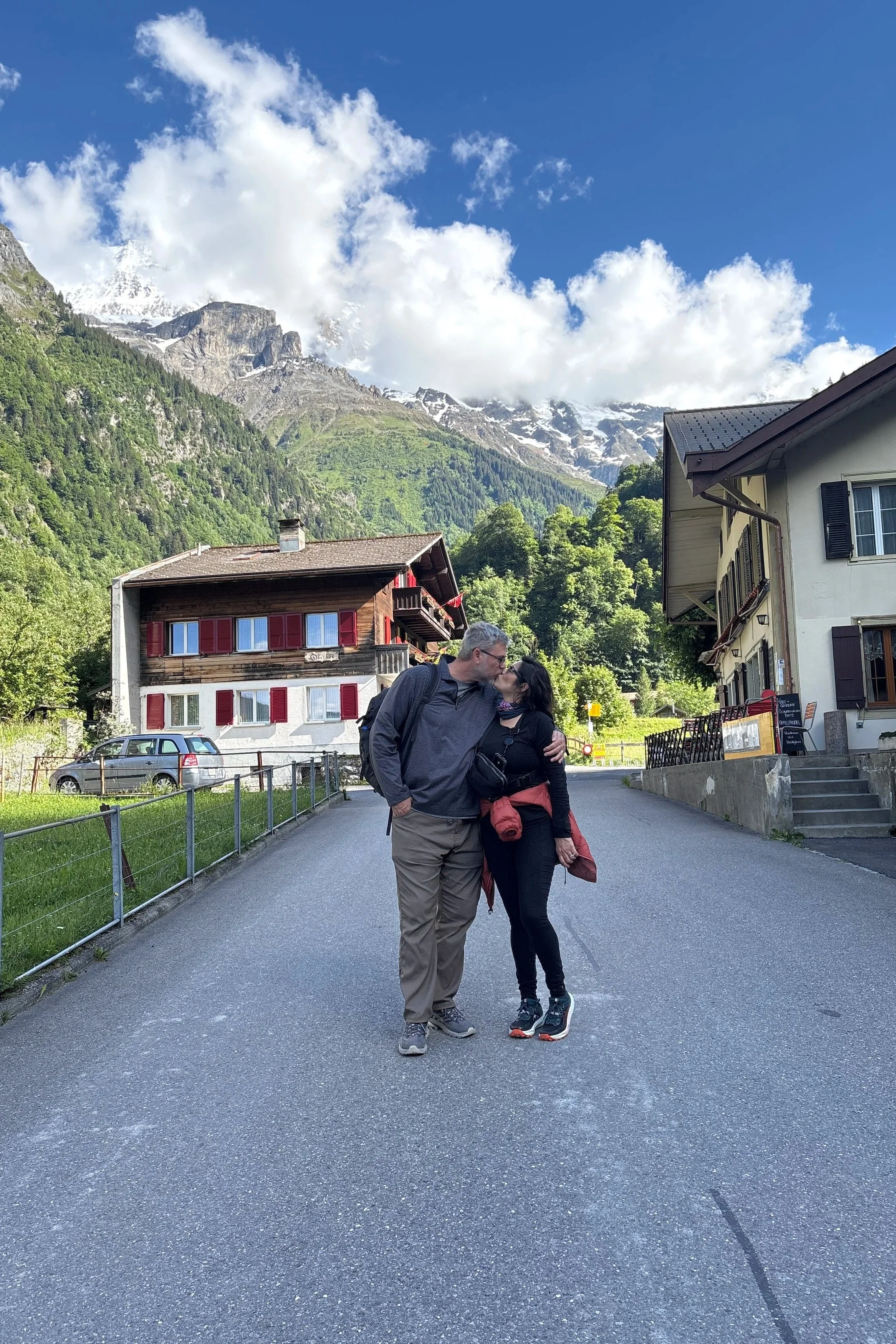 A couple kissing in a small town with mountains in the background. The man has gray hair, wears glasses, a dark jacket, and khaki pants. The woman has dark hair, wears a black jacket, and sneakers. There are houses with colorful shutters, parked cars, and lush green trees.