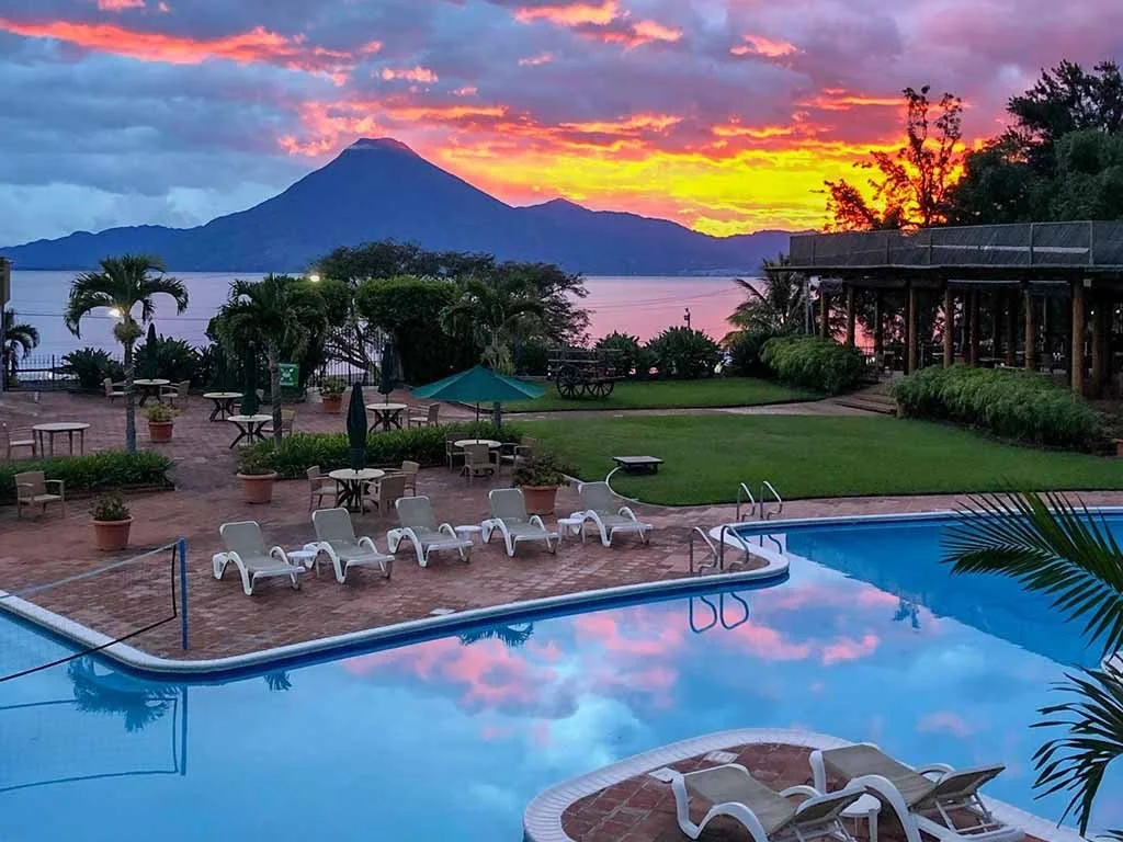 A scenic view of a pool area at sunset, with mountains and a large body of water in the background, surrounded by lounge chairs, potted plants, and a wooden building.