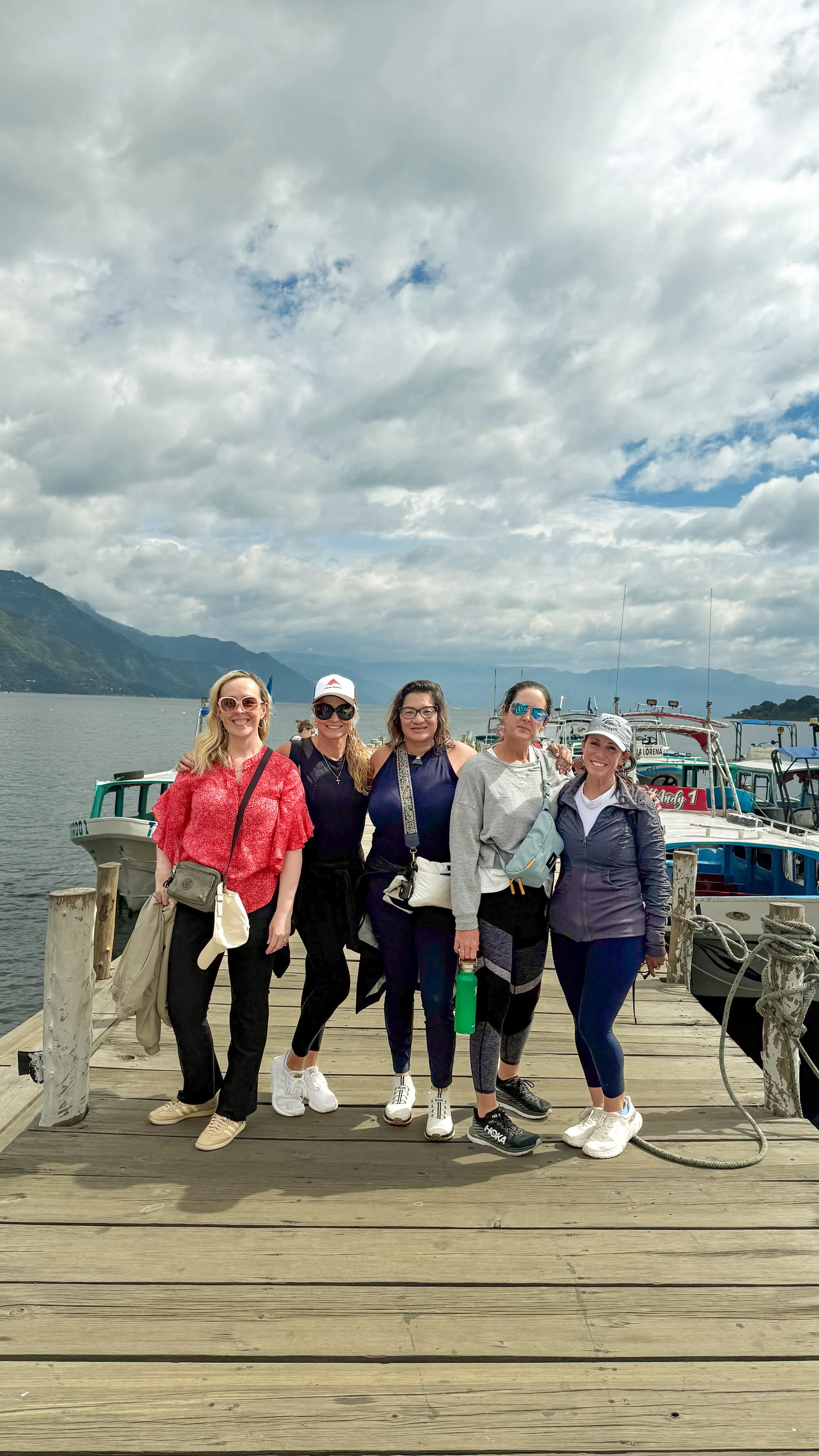 Five women standing on a wooden dock in front of boats and a lake with mountains in the background, under a cloudy sky.