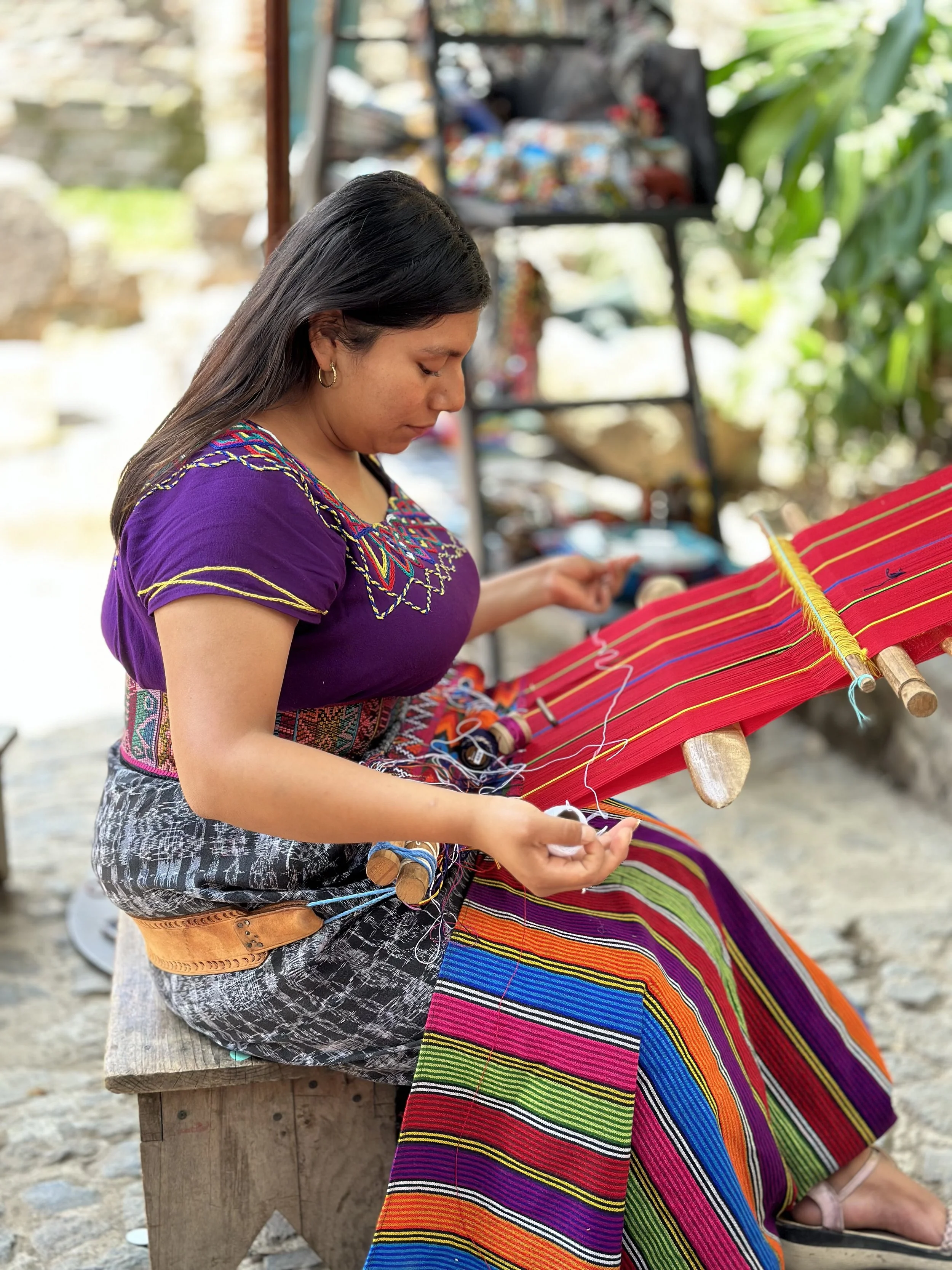 A woman sitting outdoors on a wooden bench, working on traditional colorful weaving with a loom. She is wearing a purple embroidered blouse and patterned skirt, with a focus on her craft. In the background, there are shelves with various supplies and lush green foliage.