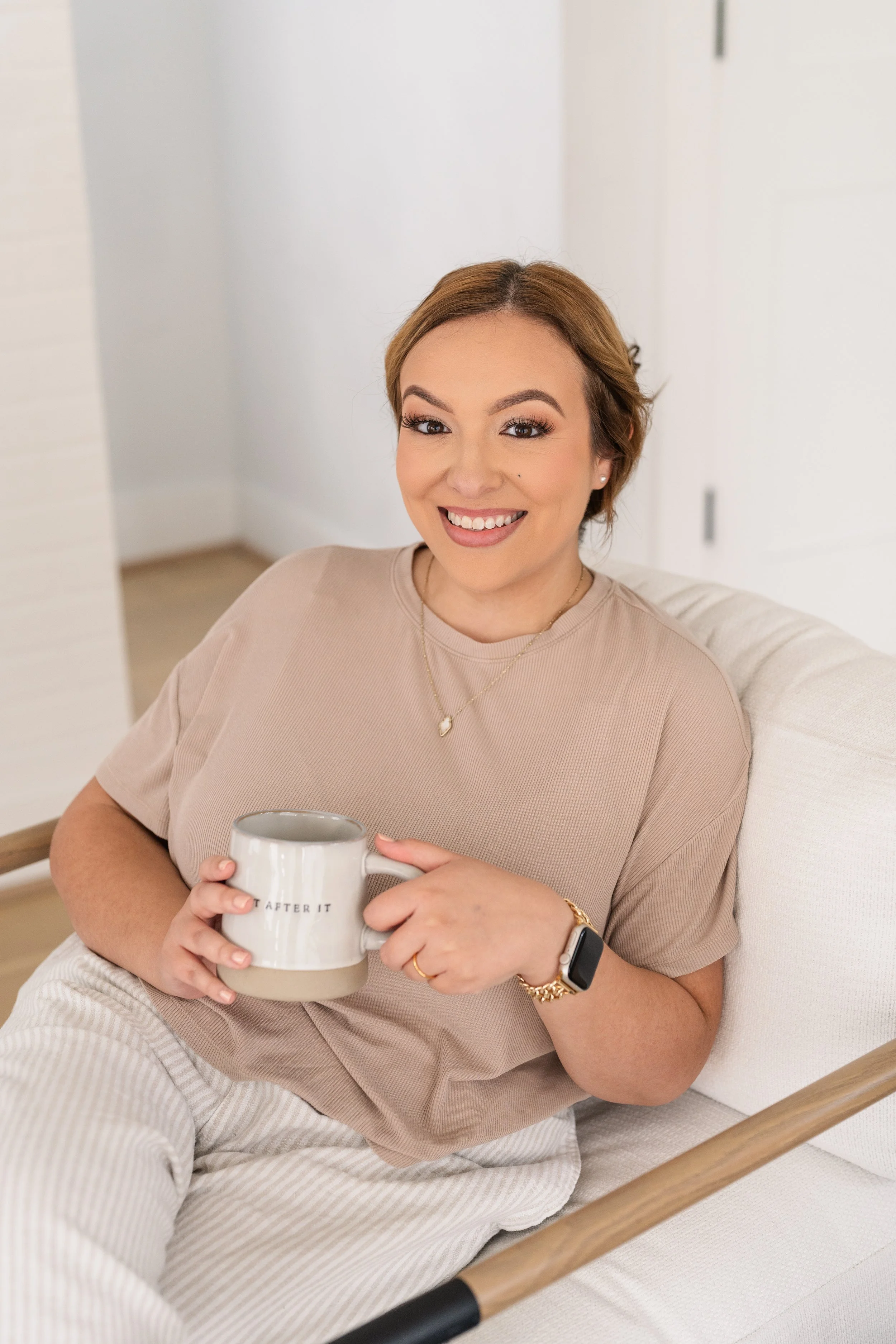 A woman with light brown hair, smiling, sitting on a light-colored sofa, holding a white mug that reads 'AFTER IT', wearing a beige t-shirt, striped pants, a watch, and jewelry in a bright, modern room.