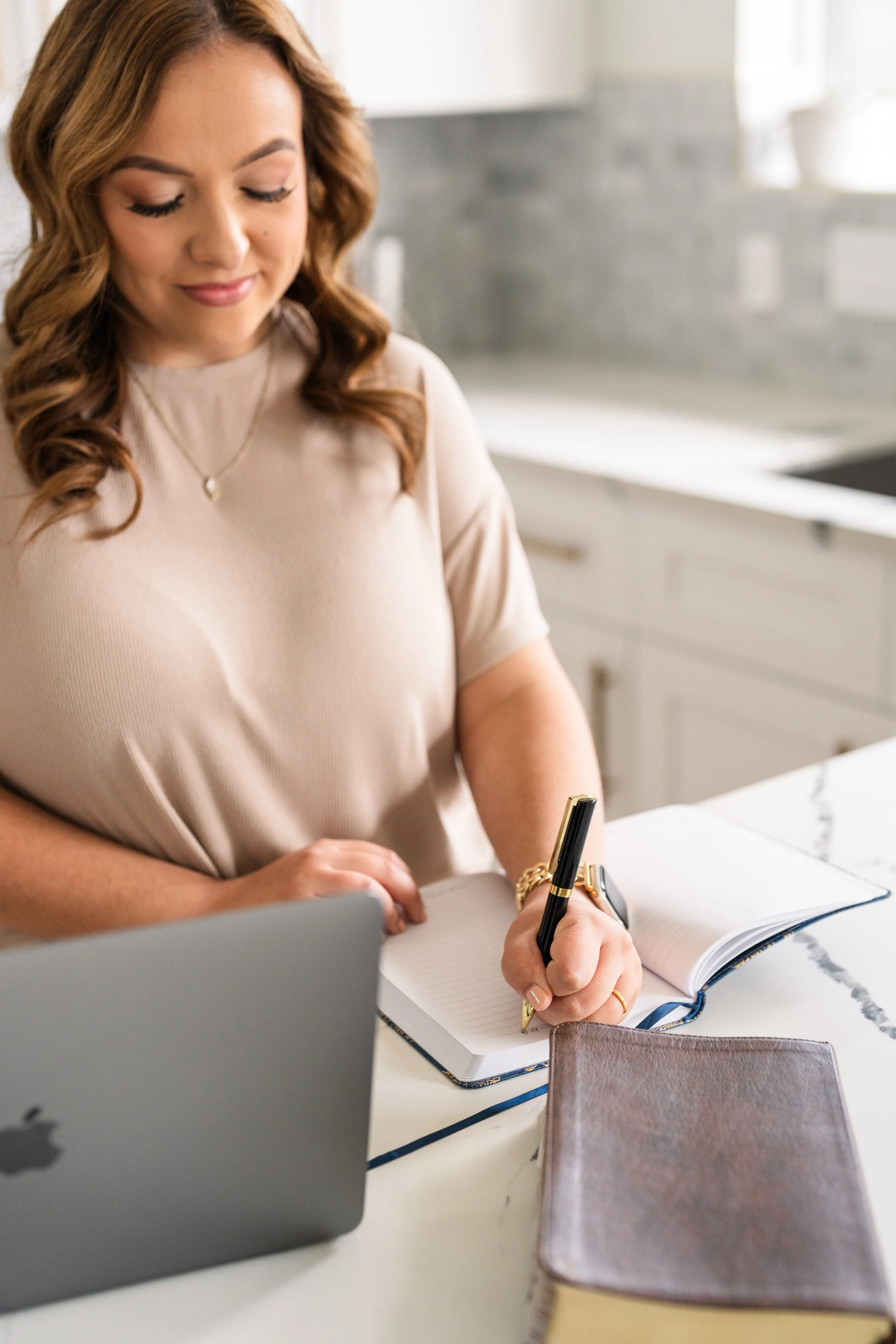 Woman writing in a notebook at a marble kitchen counter with a laptop and closed book nearby.