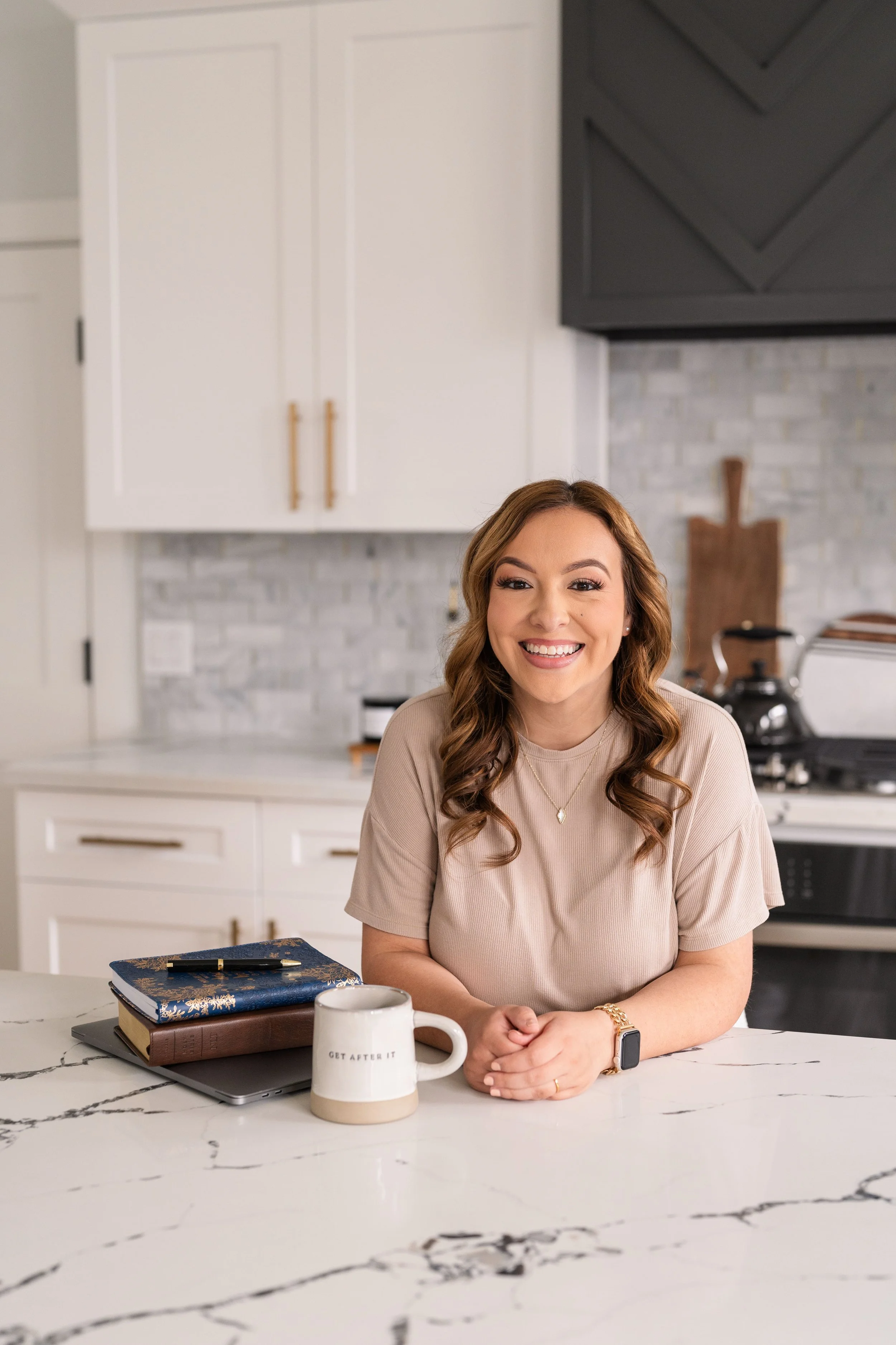 A woman smiling and sitting at a kitchen counter with a coffee mug, closed laptop, notebooks, and a pen.