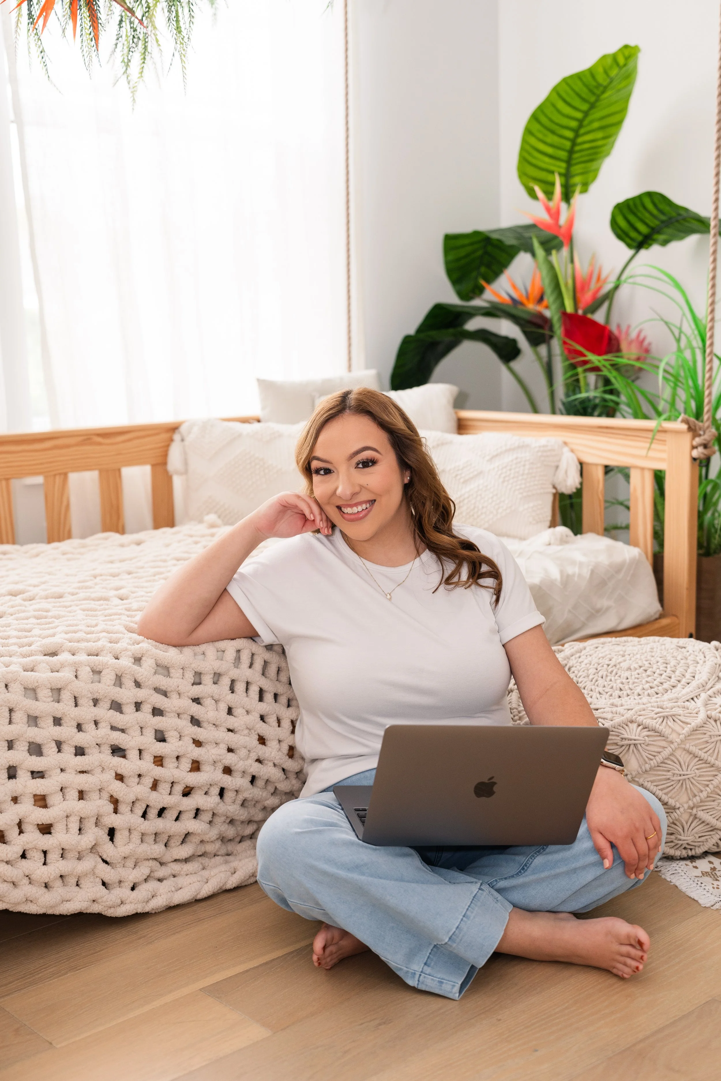 A woman sitting on the floor with a laptop, smiling, in a cozy bedroom with plants and a wooden bed frame.