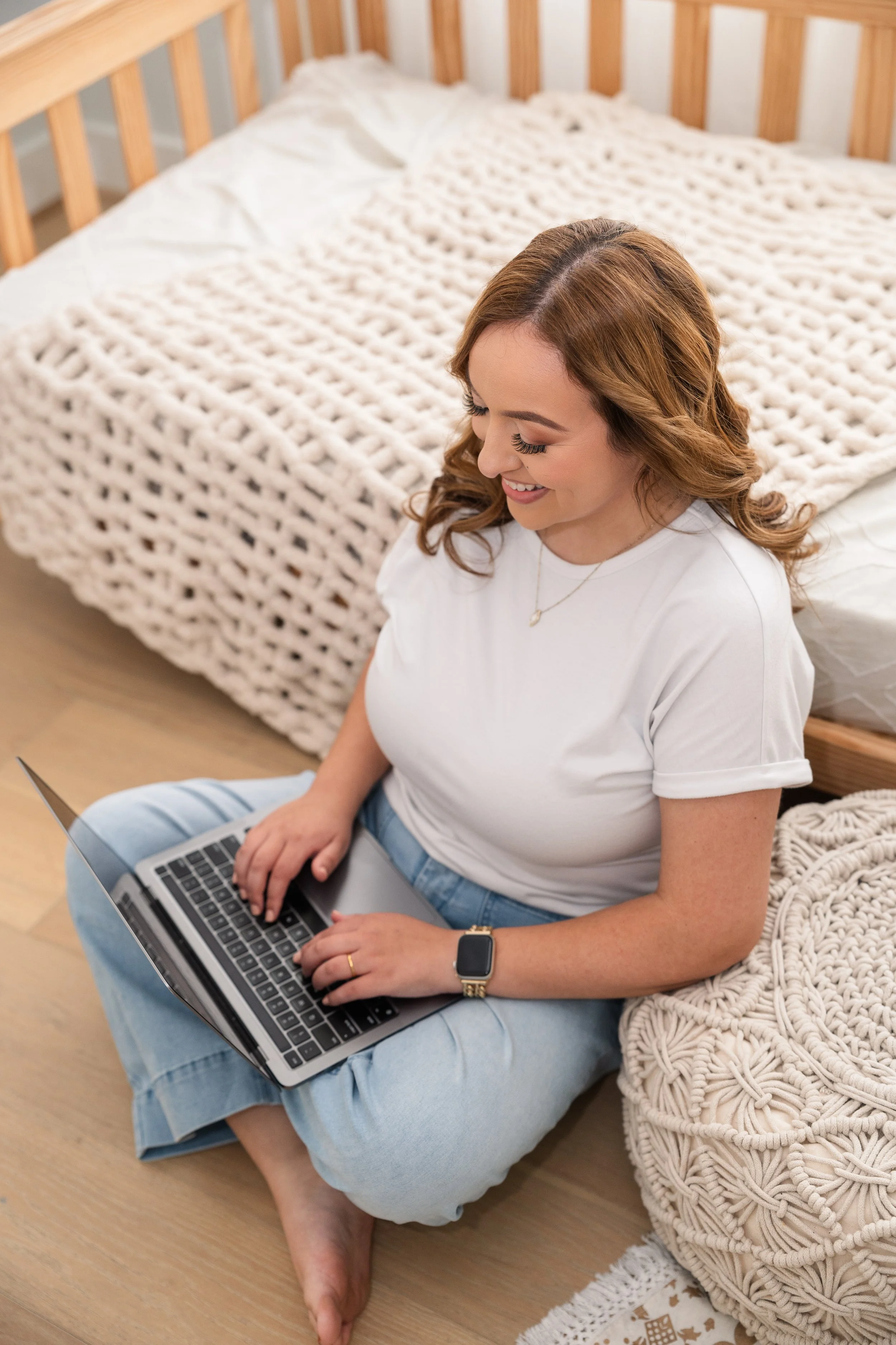 A woman sitting on the floor with her legs crossed, using a laptop near the bed in a cozy bedroom with beige knit blankets and wooden furniture.