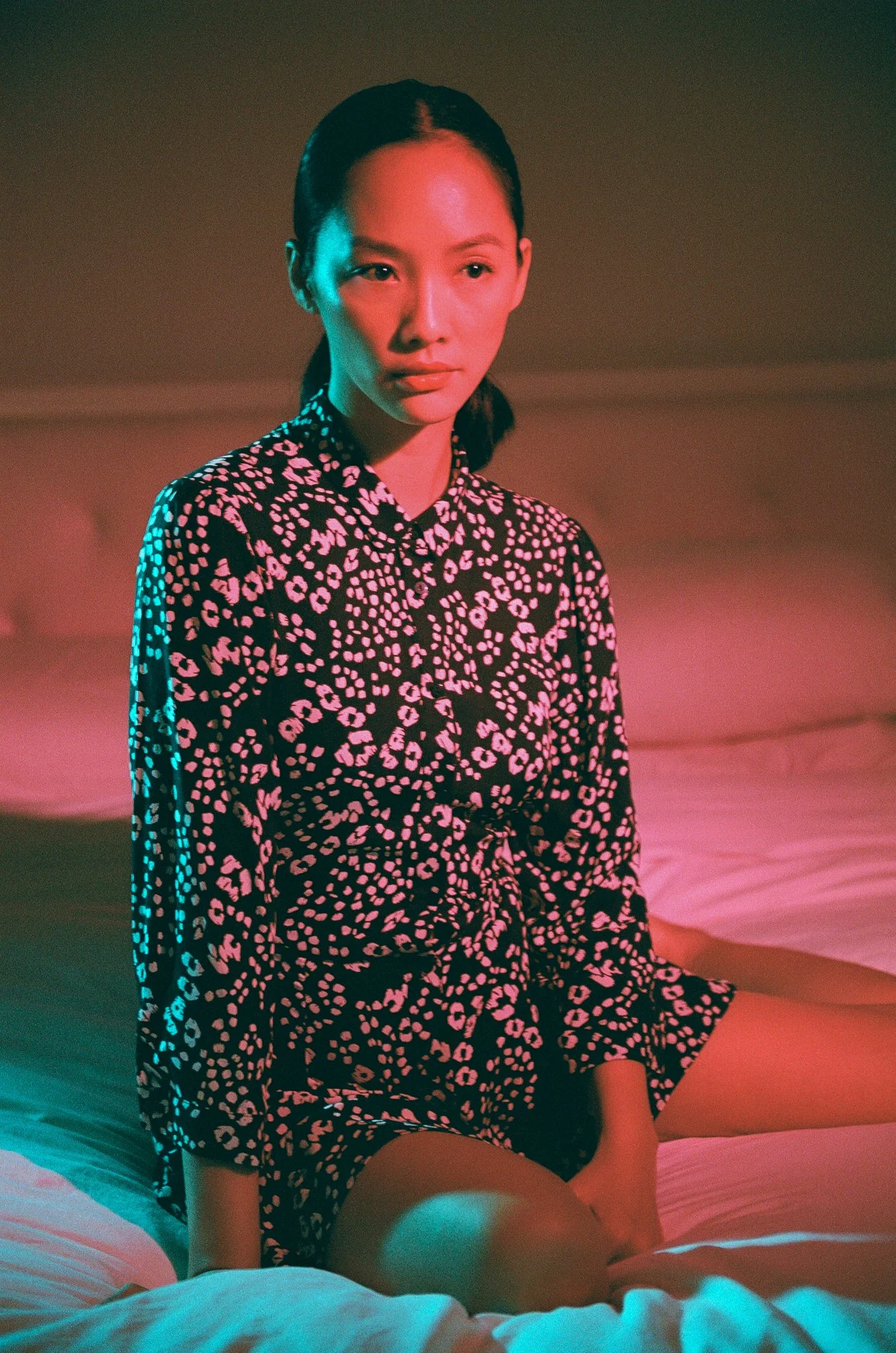 A woman sitting on a bed with a pink and black patterned dress, in a room lit with pink and green neon lights.