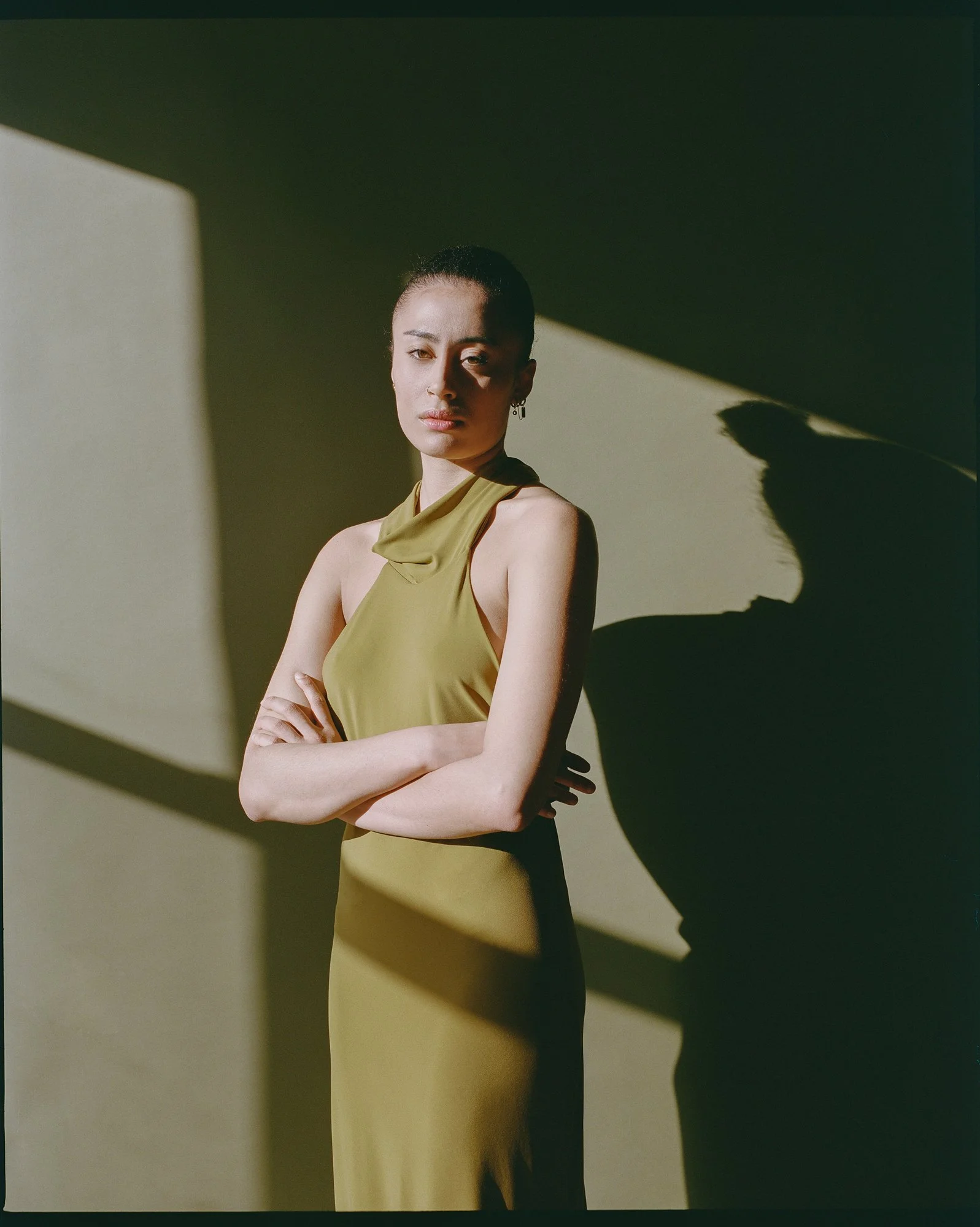 A woman with dark hair tied back, wearing a sleeveless olive green dress, stands with her arms crossed, in front of a shadow cast on a yellowish-green wall.