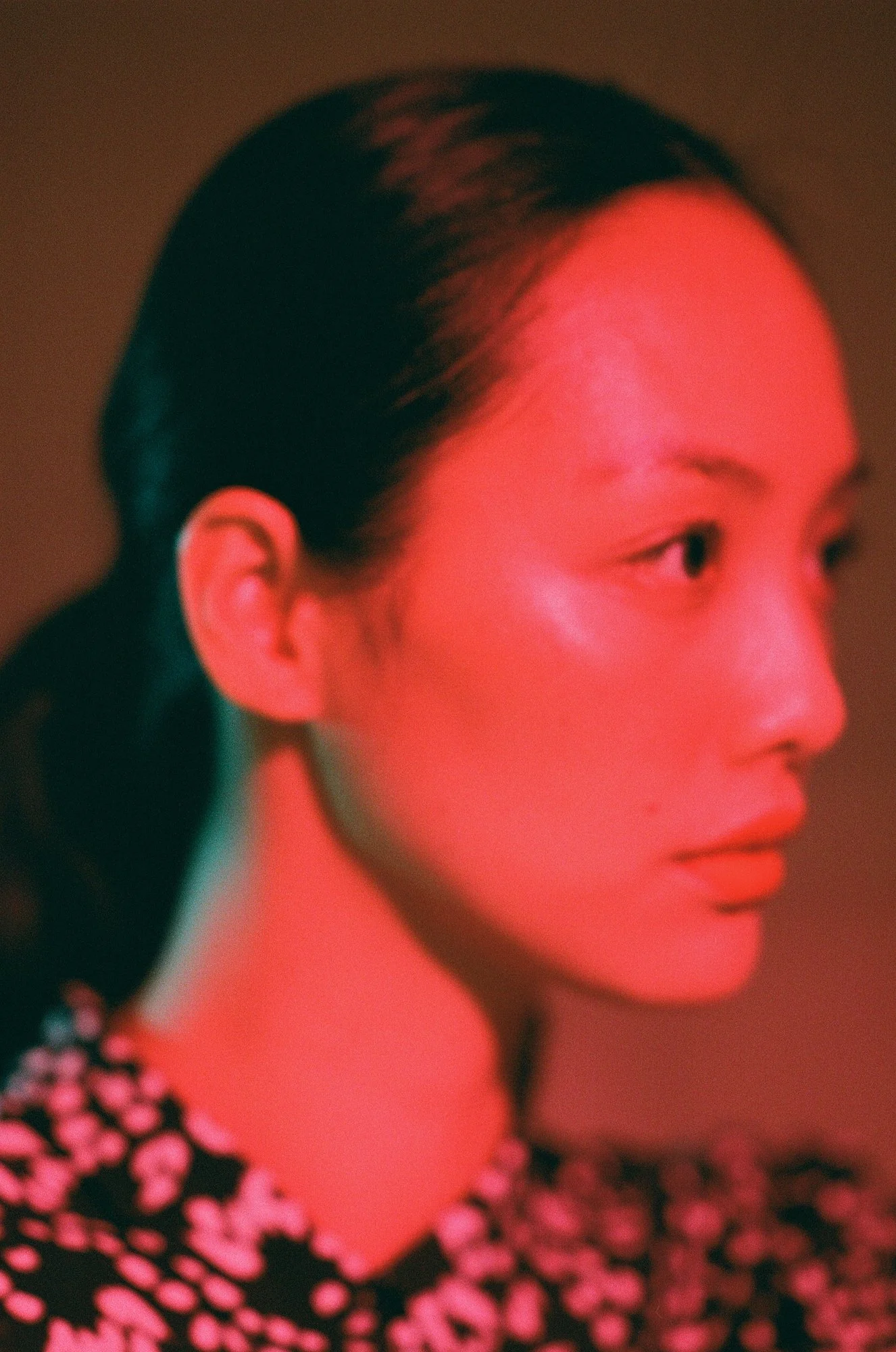 Close-up side profile of an Asian woman with dark hair, wearing a black dress with pink floral pattern, illuminated with red and pink lighting.