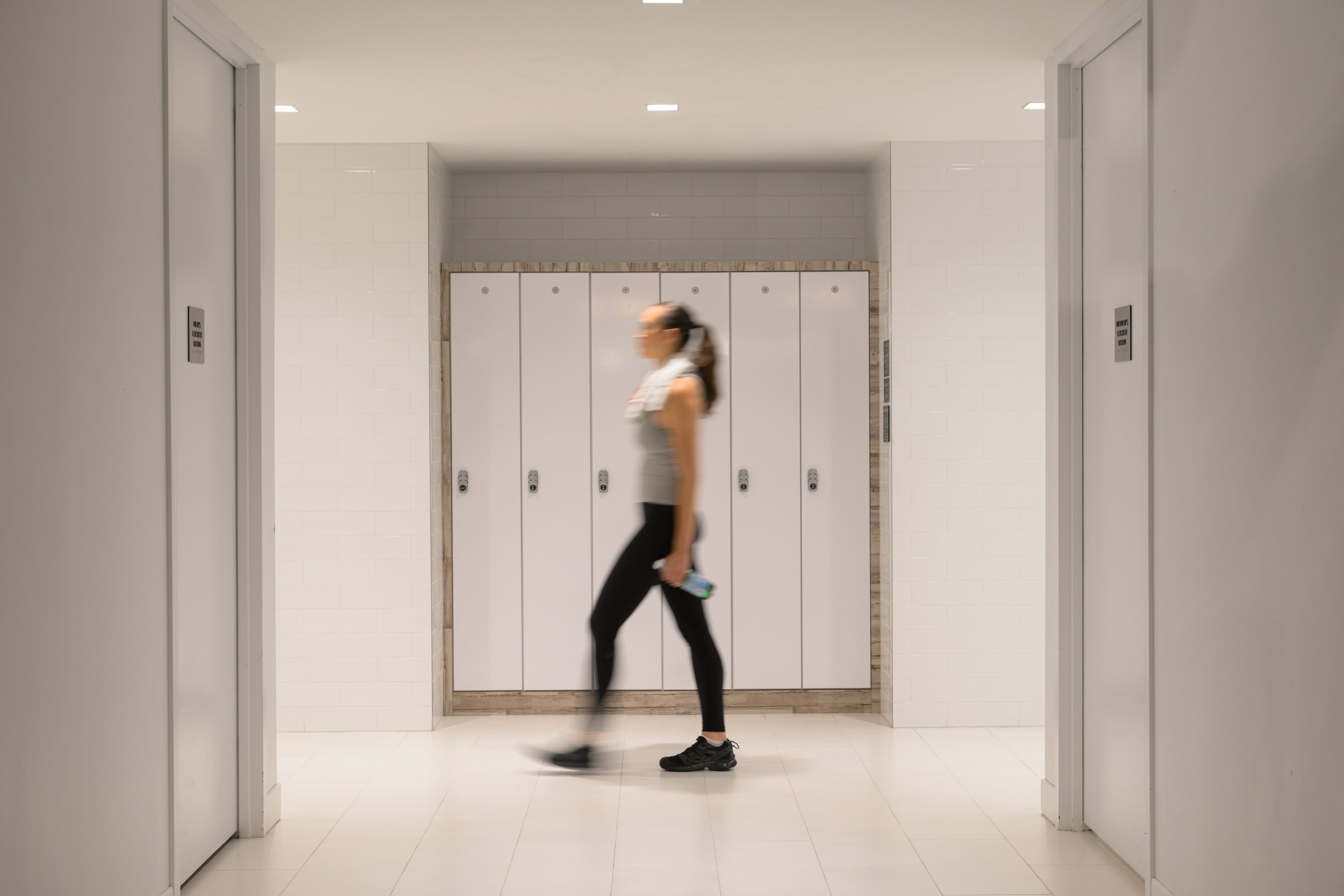 A woman walking past bathroom stalls in a modern, well-lit hallway.