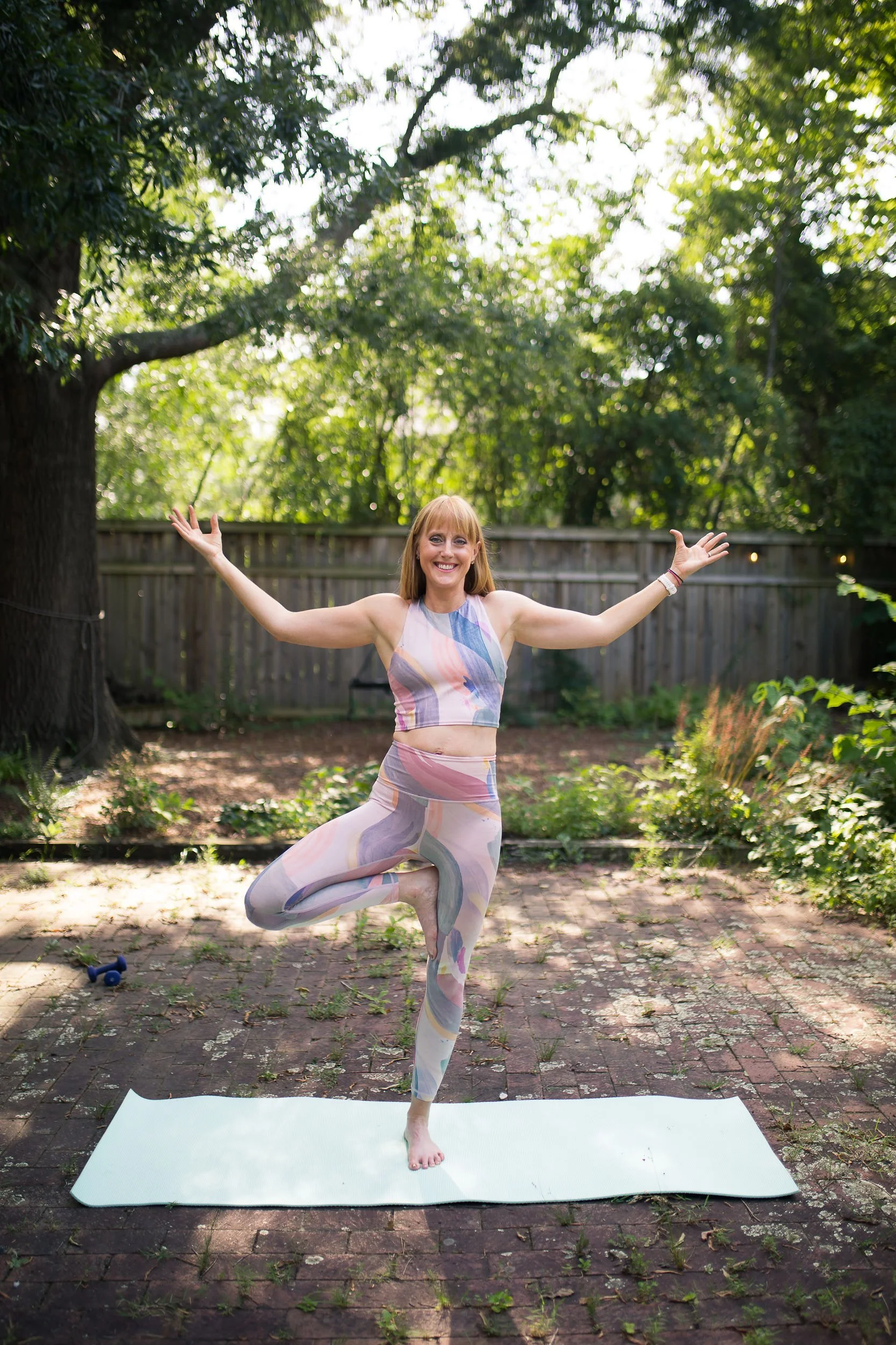 A woman practicing yoga outdoors on a yoga mat in a garden with green trees and a wooden fence, smiling and balancing on one leg with arms stretched out.