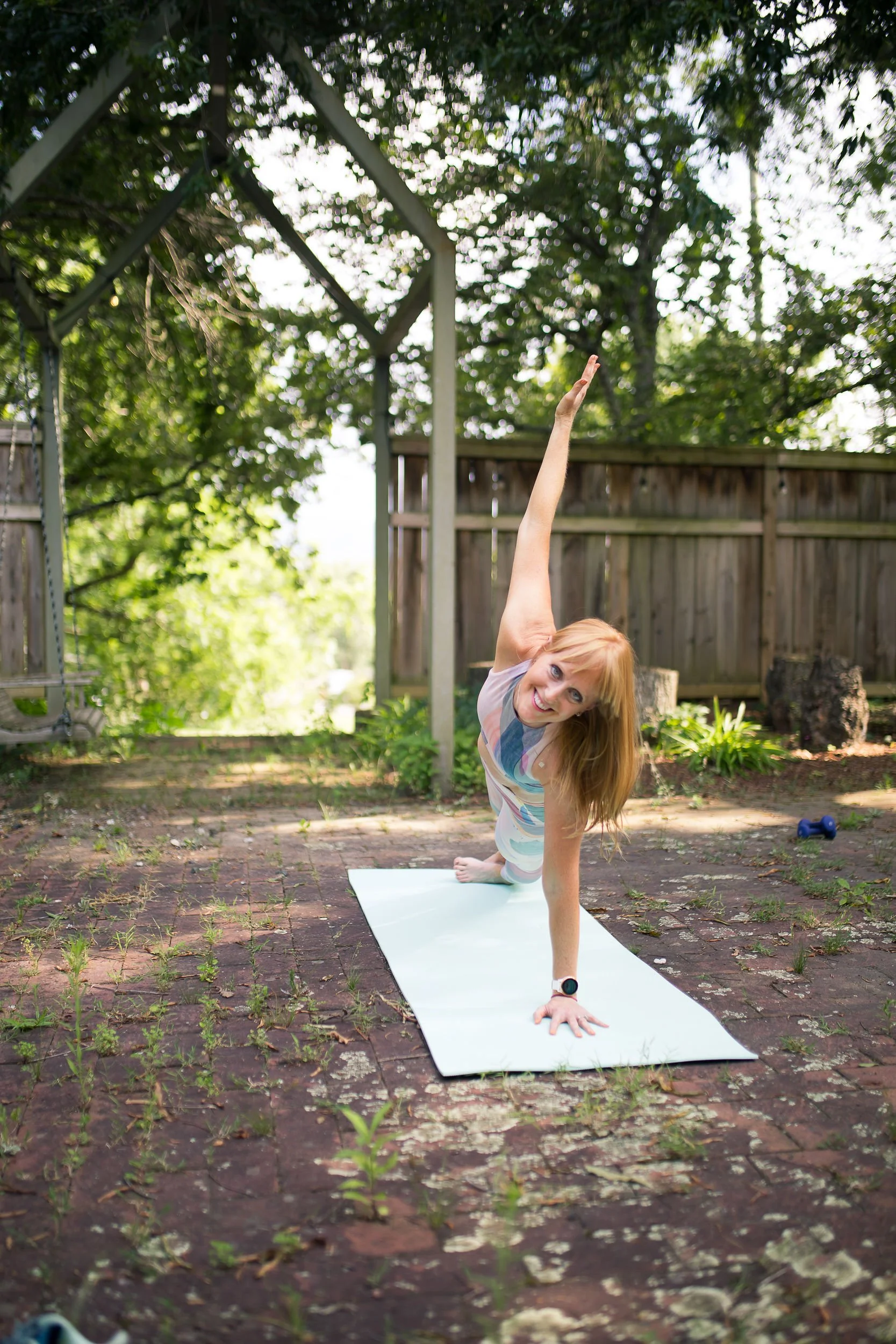 Woman doing a yoga pose on a white mat outdoors, surrounded by trees and a wooden fence, with a smiling expression.