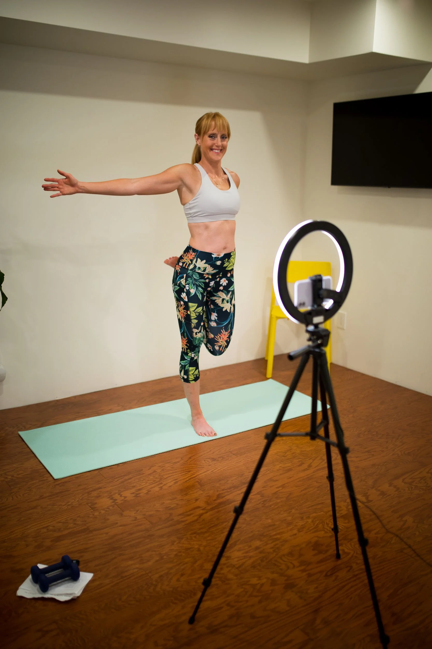 Woman in workout attire practicing a yoga pose in front of a camera and ring light in a room with hardwood floors.