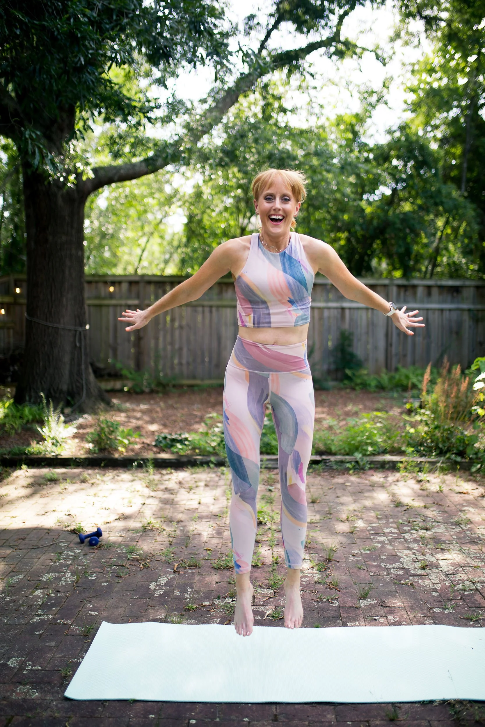 A woman in colorful athletic wear jumps in the air on a yoga mat outdoors, smiling joyfully. She is surrounded by trees and a wooden fence, with a brick patio under her.