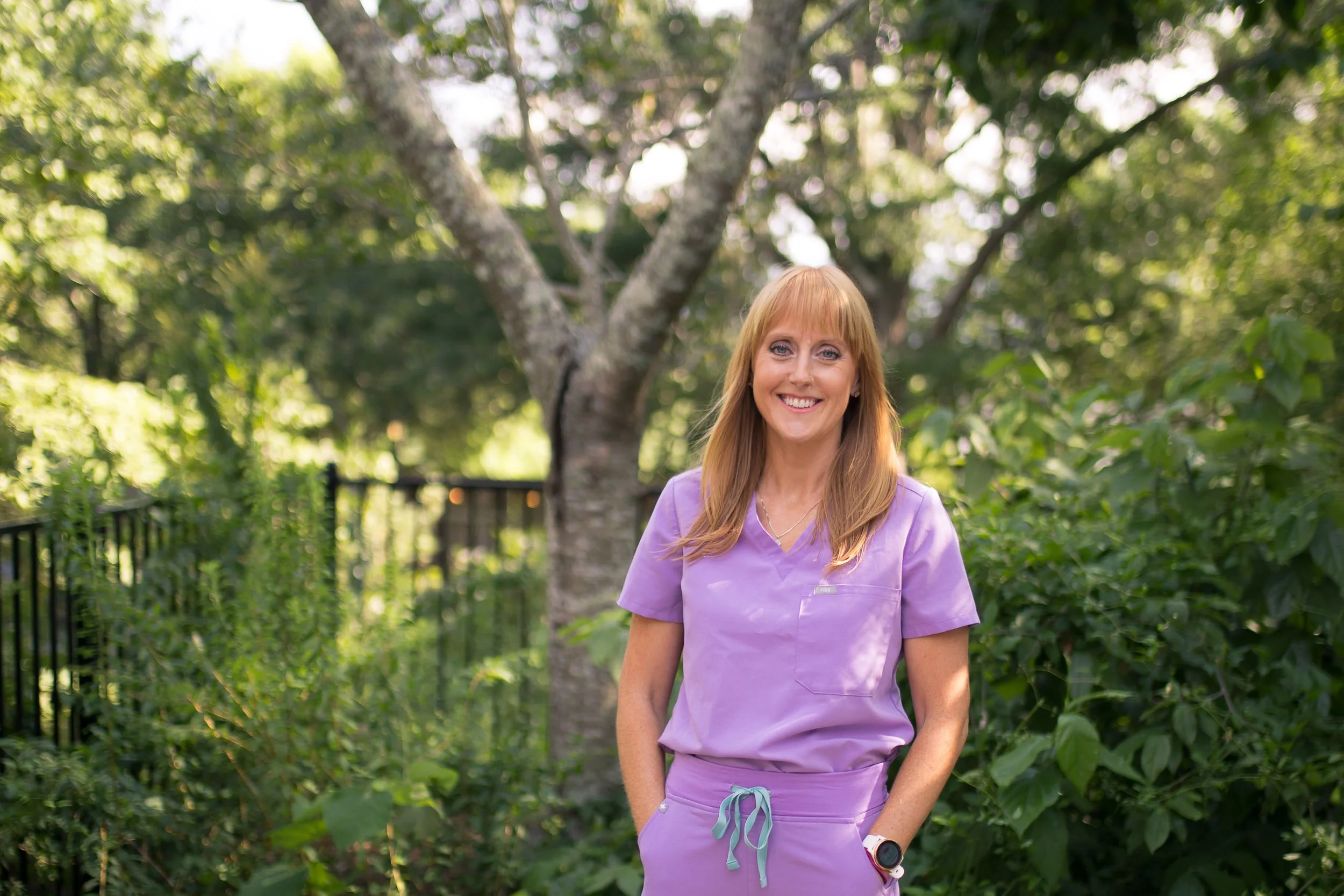 A woman in purple scrubs standing outdoors in a lush green garden with a large tree behind her, smiling at the camera.