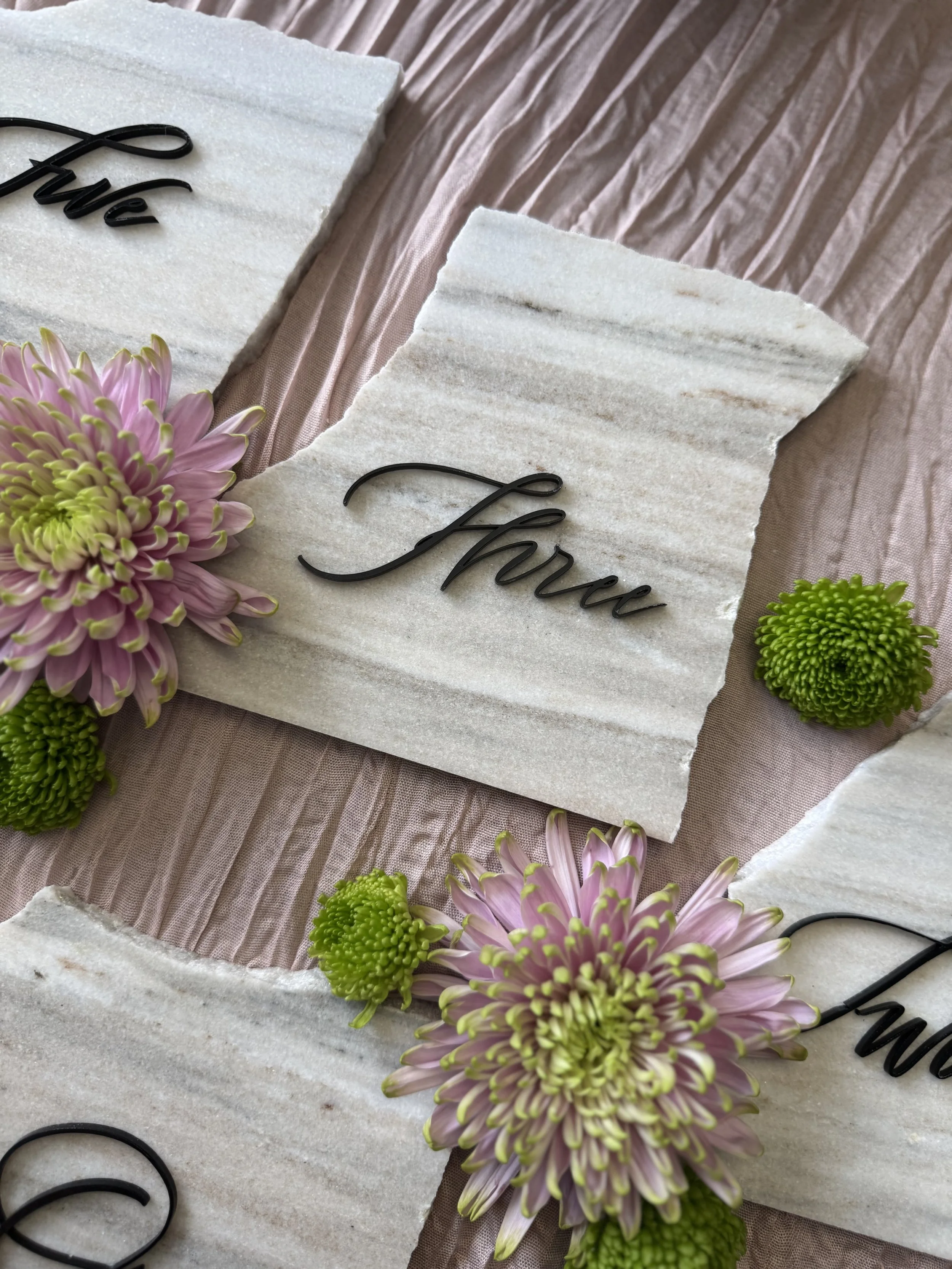 A decorative arrangement featuring pink and purple flowers, green moss balls, and sections of white and beige textured paper or fabric with black cursive words.