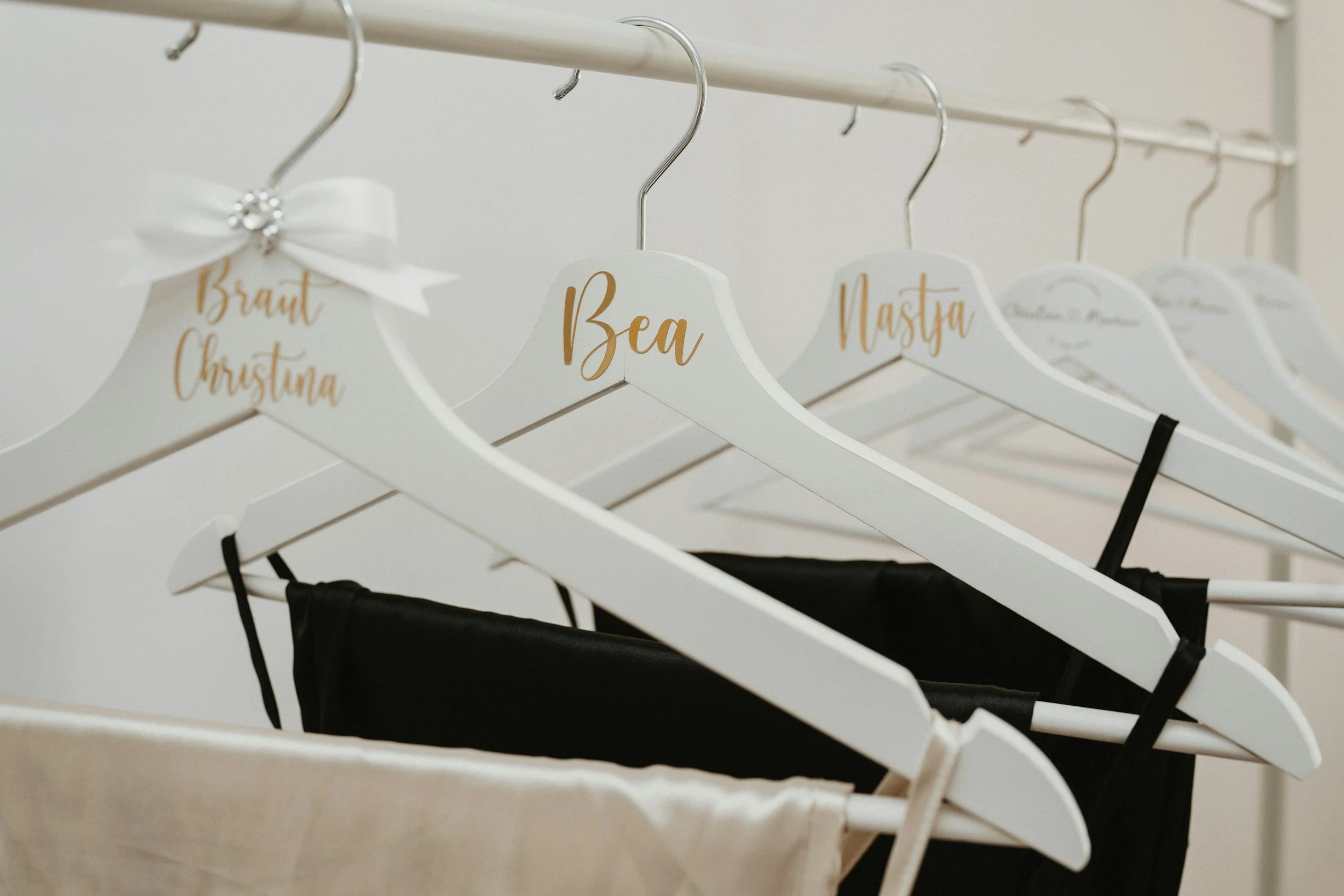 A row of white clothing hangers on a metal rack, with names written in gold font, including 'Bea' and 'Brant Christina'. Some hangers have black clothing items, and one hanger is decorated with a white bow and rhinestones.