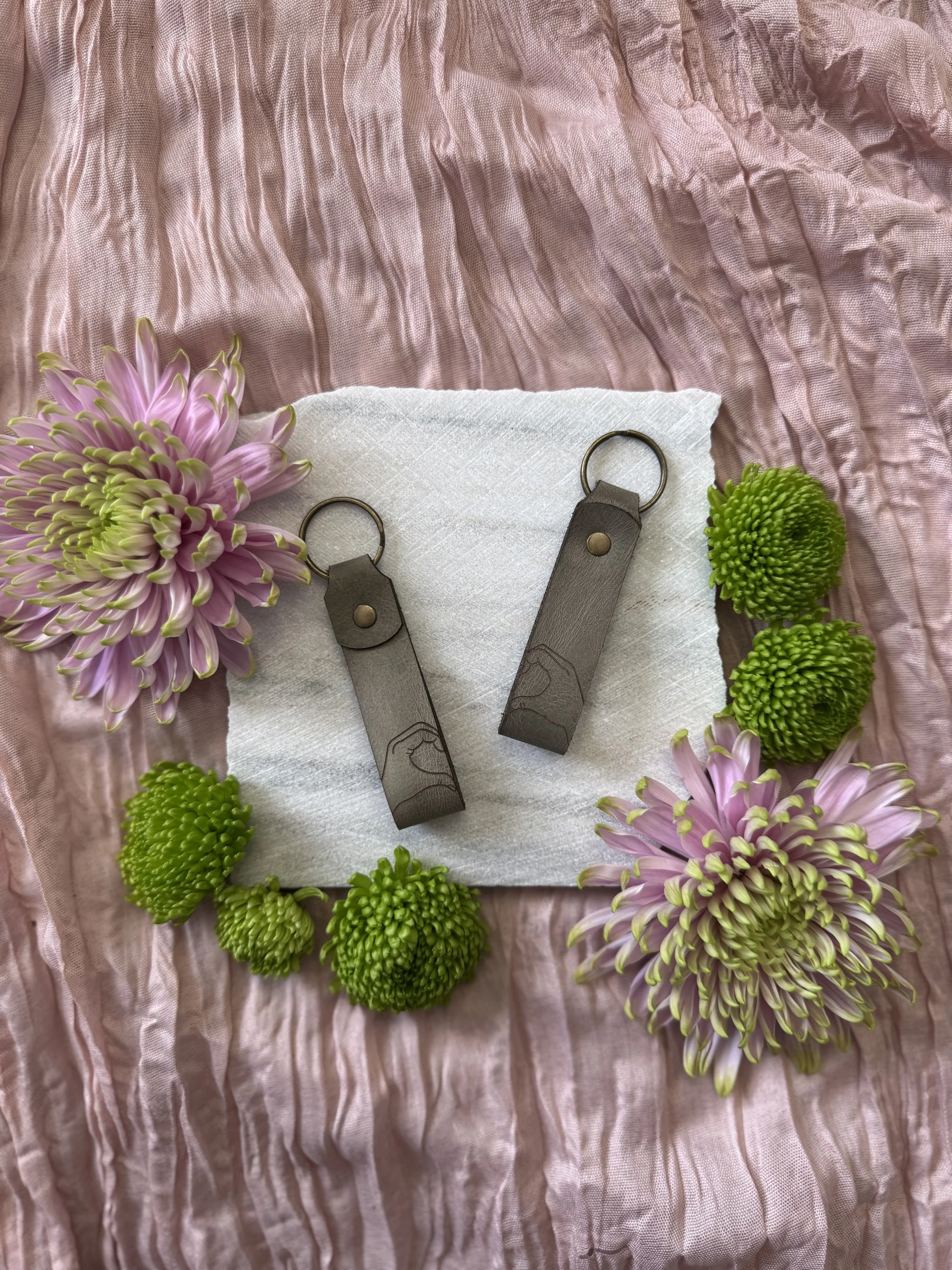 Decorative setup with pink flowers, green floral foam, and leather keychains on white fabric, all on a wrinkled pink satin background.