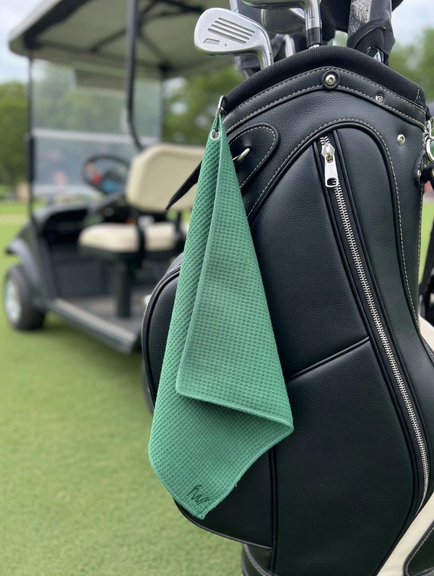 Close-up of a black golf bag with green towel hanging on it, positioned on a golf cart on a golf course.