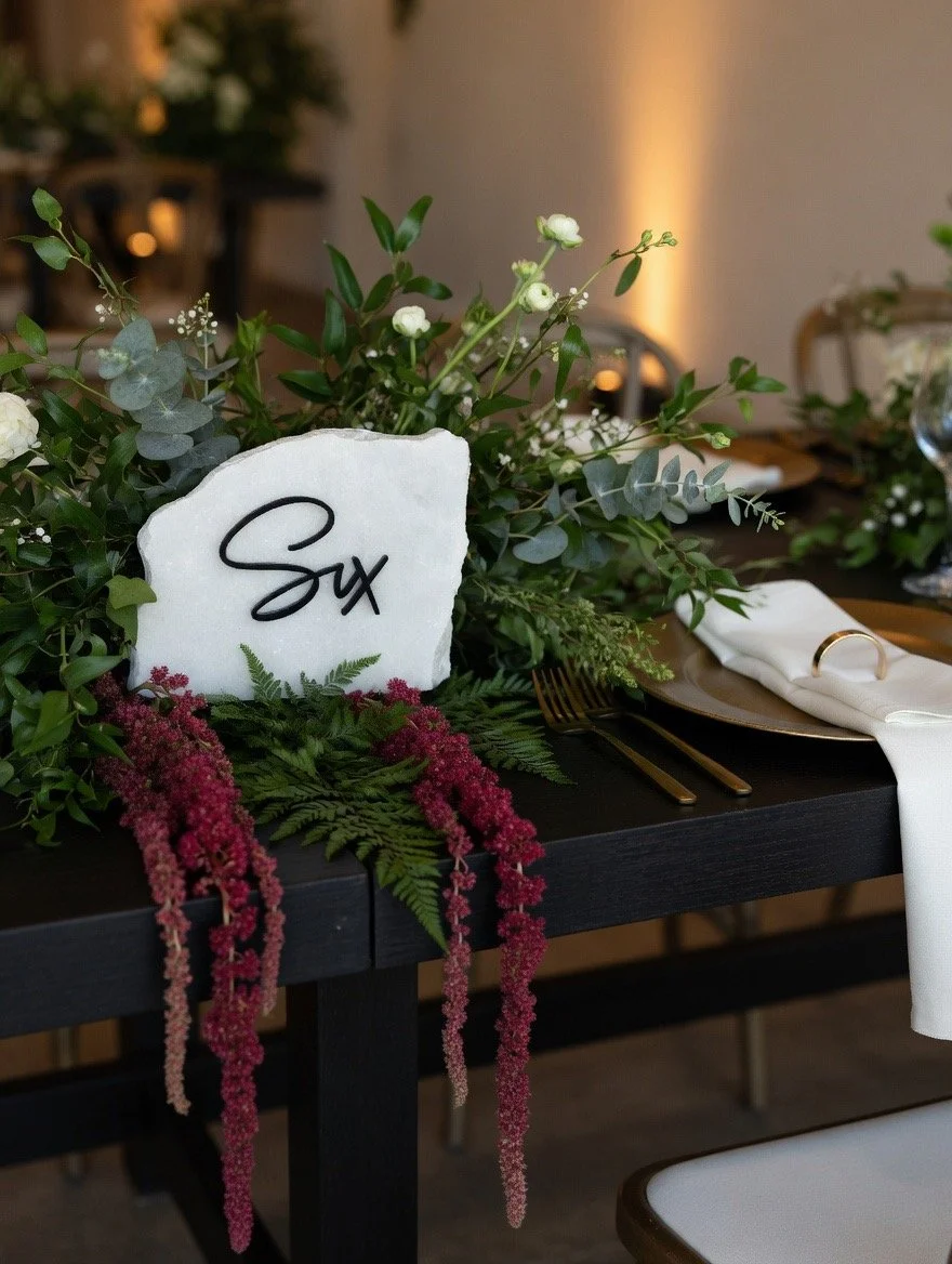 Table centerpiece with green foliage, white flowers, and burgundy hanging blooms, with a white stone displaying the name 'Six' in black cursive writing, set on a black table with gold cutlery, a white napkin, and a napkin ring.