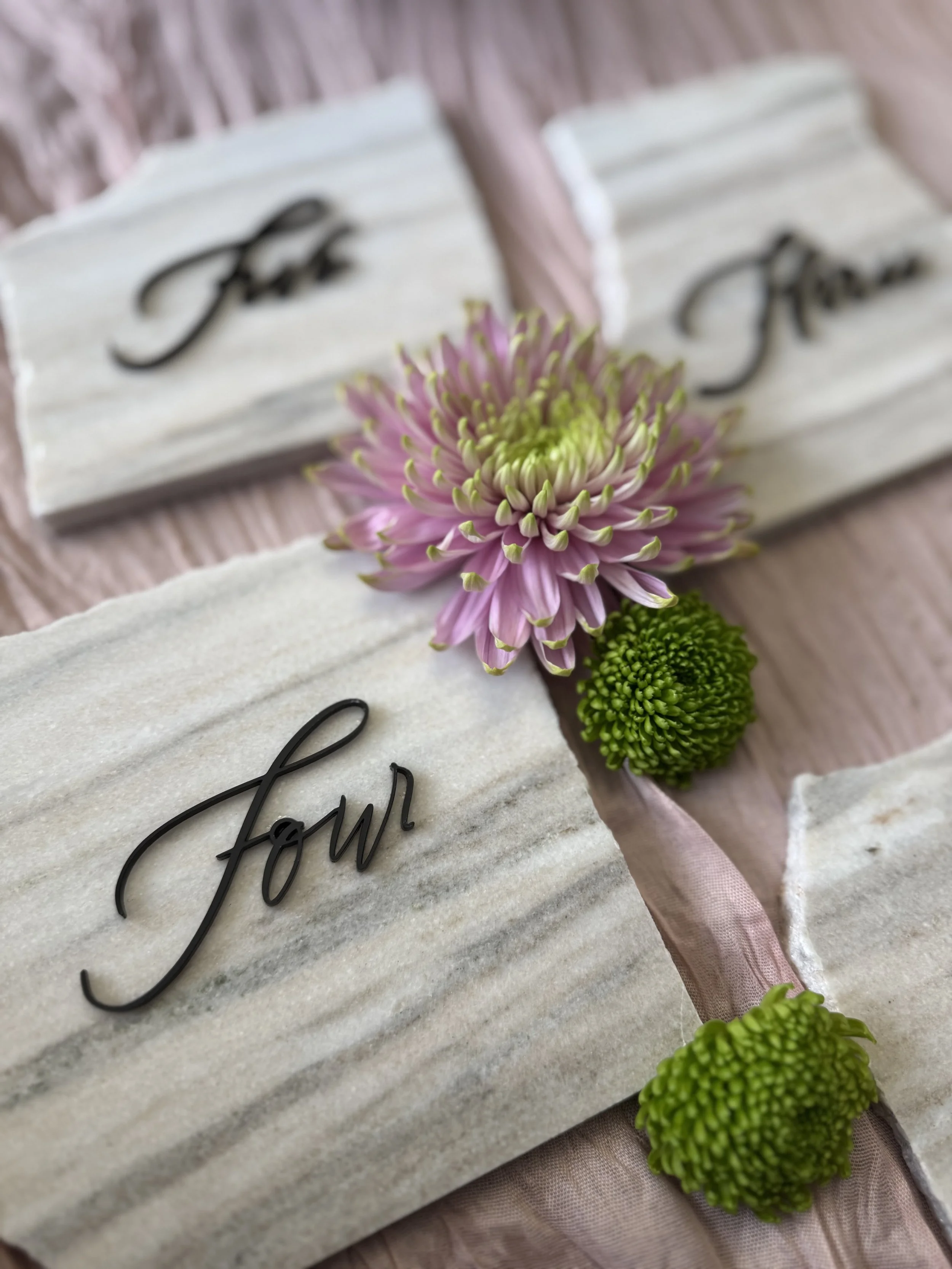 Close-up of a floral arrangement with pink and green flowers placed on a marble surface, which has black cursive writing that spells out 'for'.
