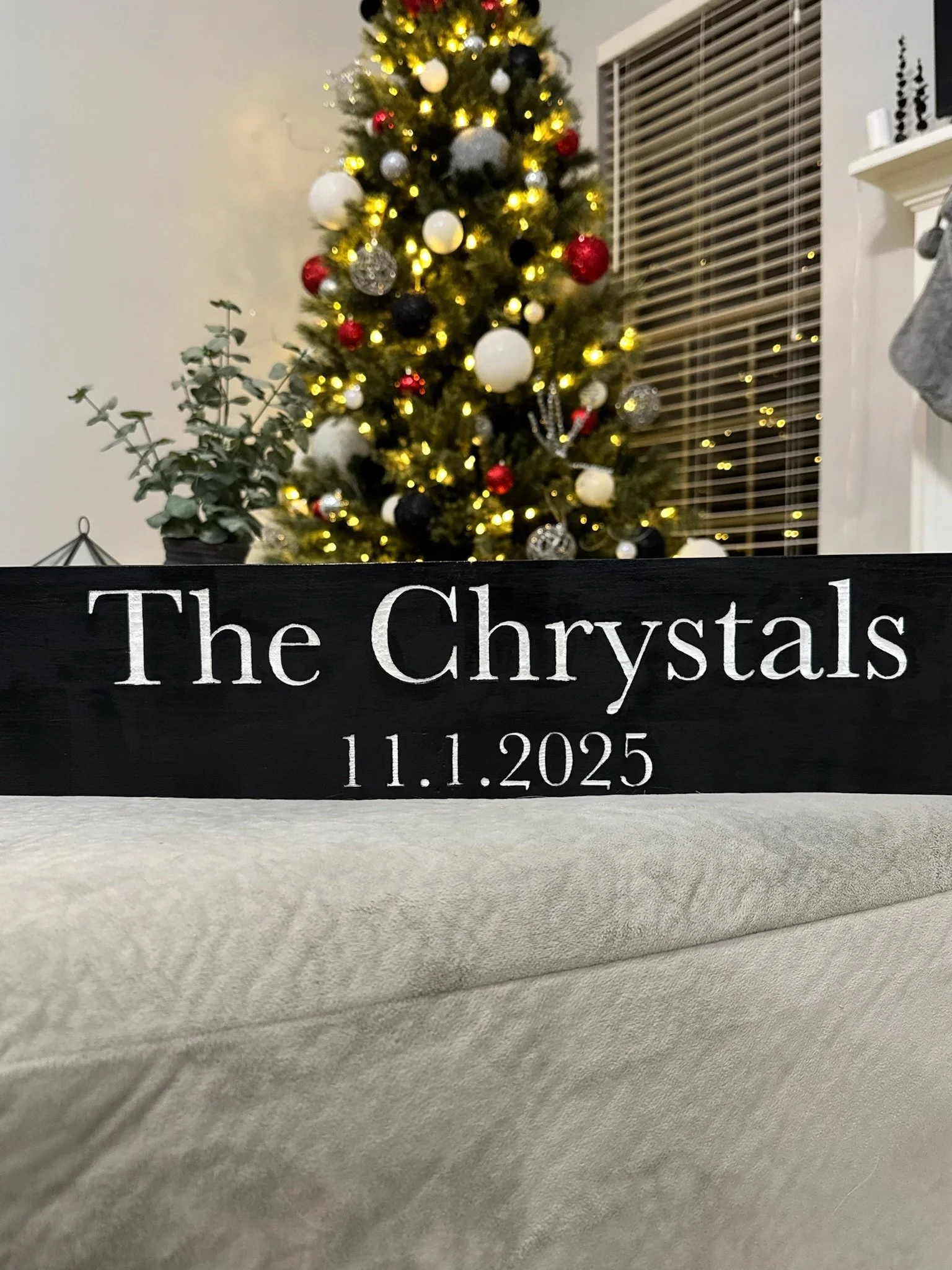 A decorated Christmas tree with white, red, black, and silver ornaments, and yellow lights, in a room with a window and a white wall. In the foreground, there is a black sign with white text that reads 'The Chrystals 11.1.2025.'