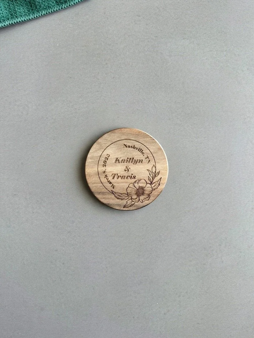 A circular wooden token engraved with floral designs and text including names 'Kaitlyn & Travis', the date 'February 2023', and the words 'Handmade'.