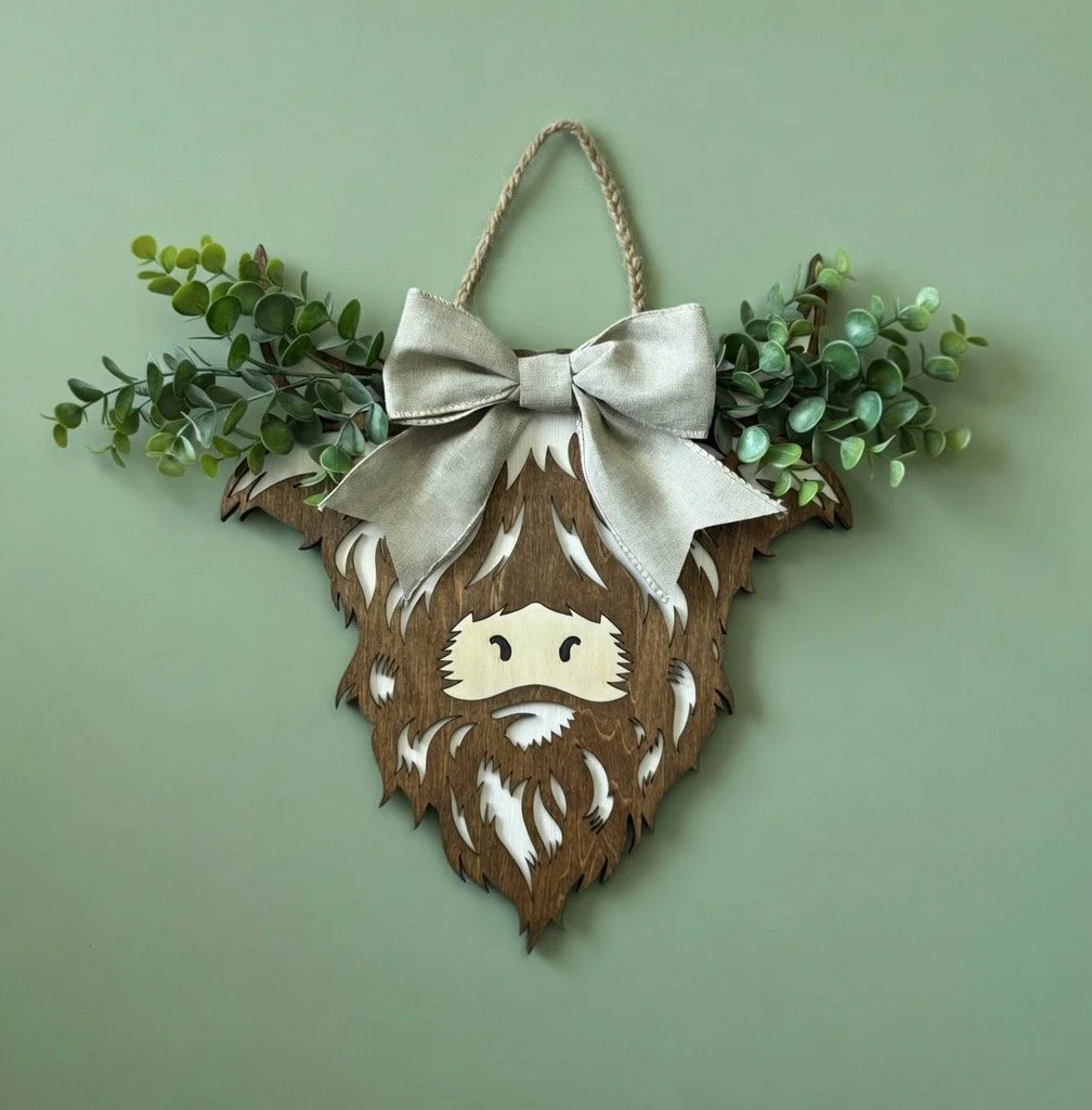 Decorative wall hanging of a wooden sheep with artificial greenery and a silver bow, attached to a green wall.