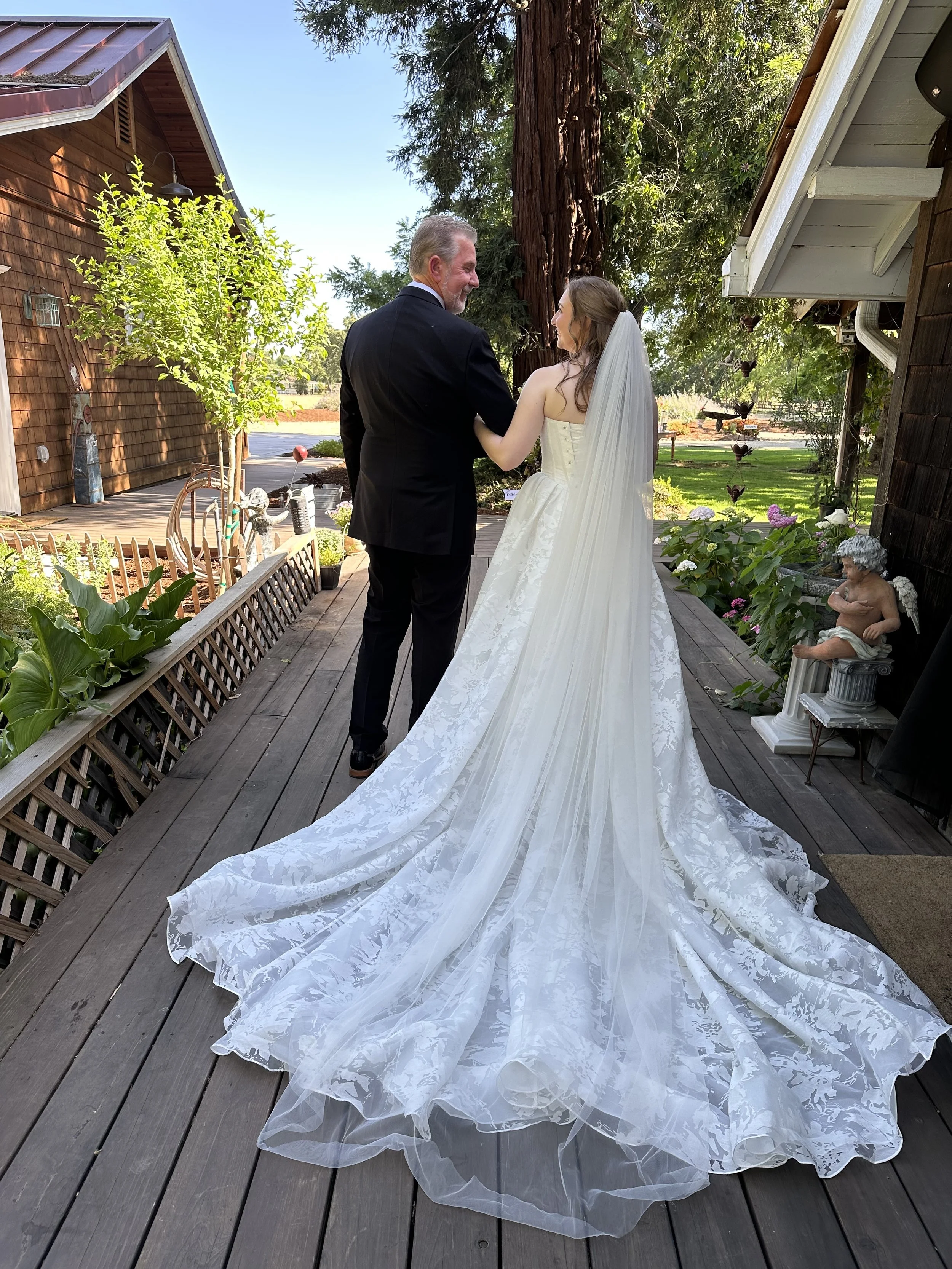 A bride and groom standing on a wooden deck outdoors, facing each other, with a large tree and plants in the background. The bride is wearing a white wedding dress with a long train and veil, and the groom is wearing a black suit. There is garden decor and a statue of a cherub on the right side of the deck.