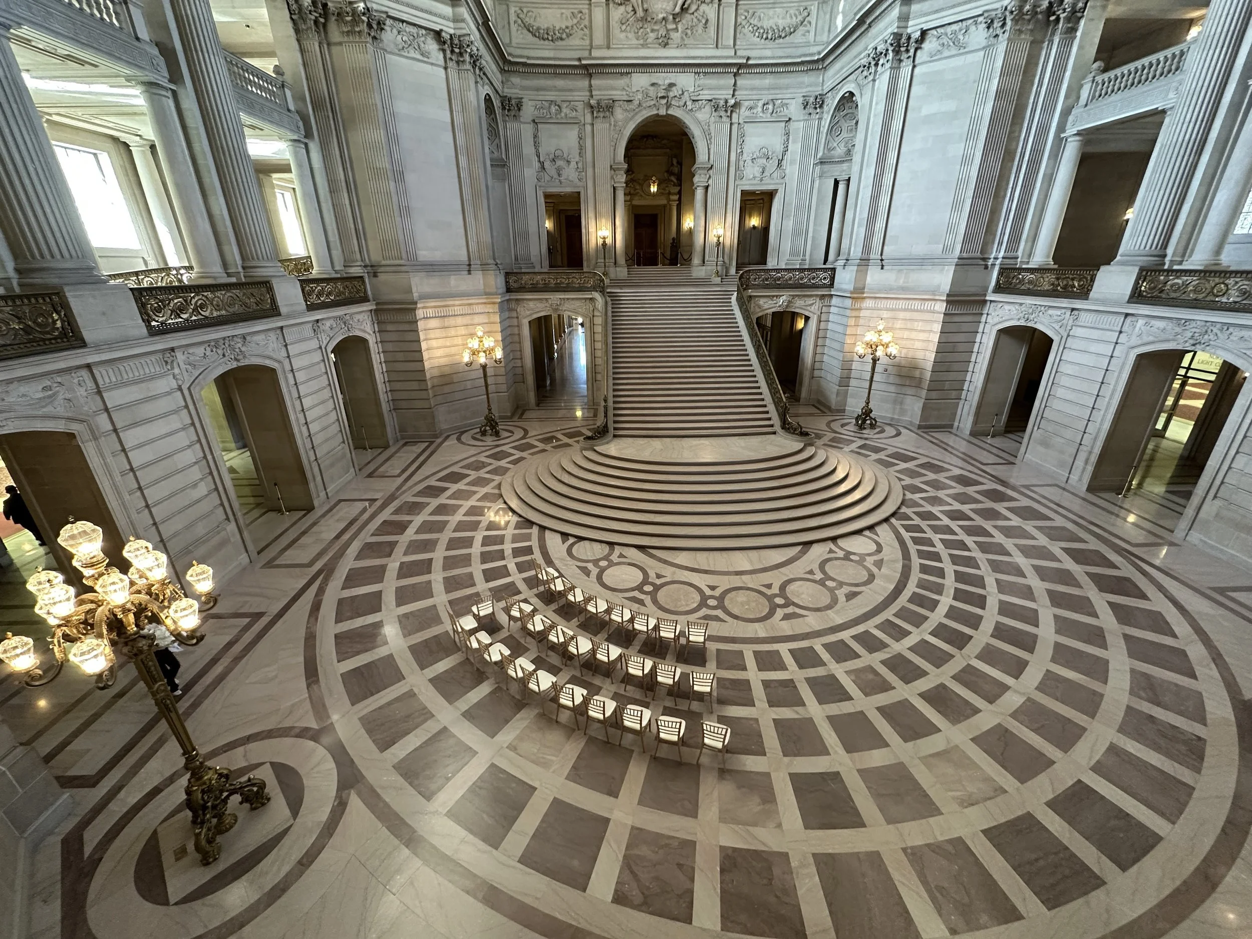 The image shows an ornate, grand staircase in a luxurious building with a large, open marble foyer. There are chandeliers hanging from the ceiling, tall windows, and decorative sculptures and moldings on the walls. The staircase is centrally located, with curved steps and detailed railings.