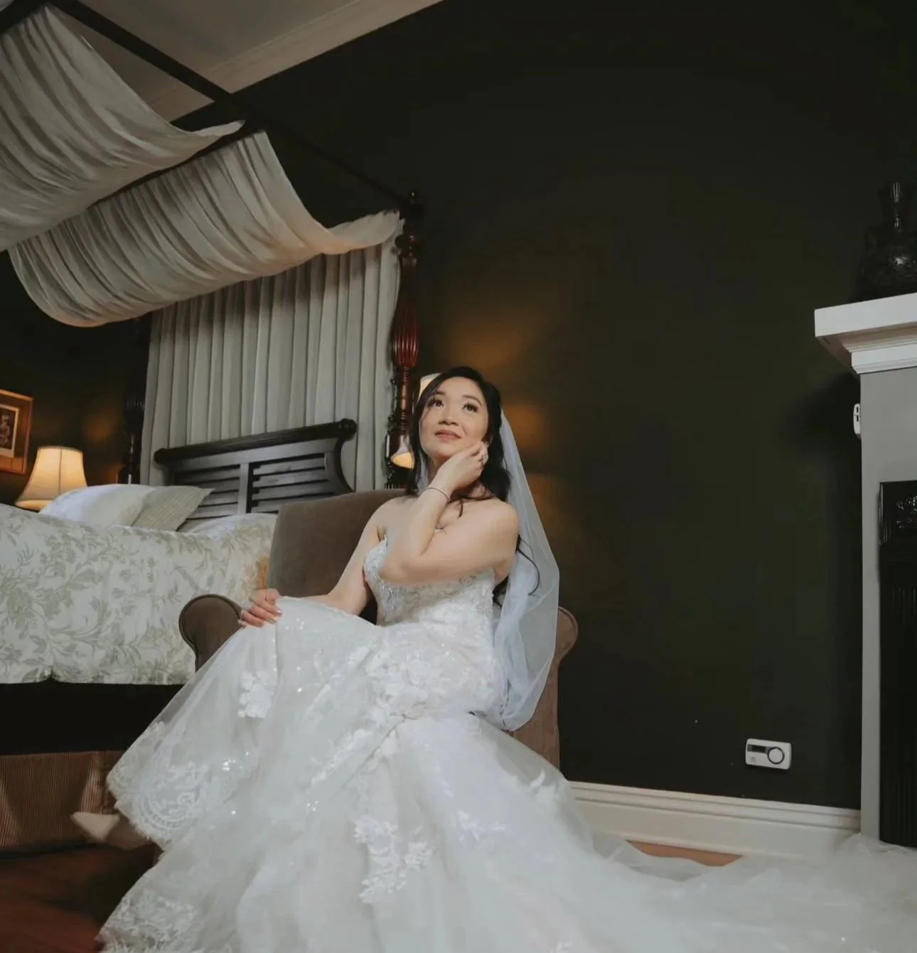 Bride sitting on a chair in a wedding dress, touching her face, in a warmly lit room with dark walls, a bed with pillows, and a fireplace.