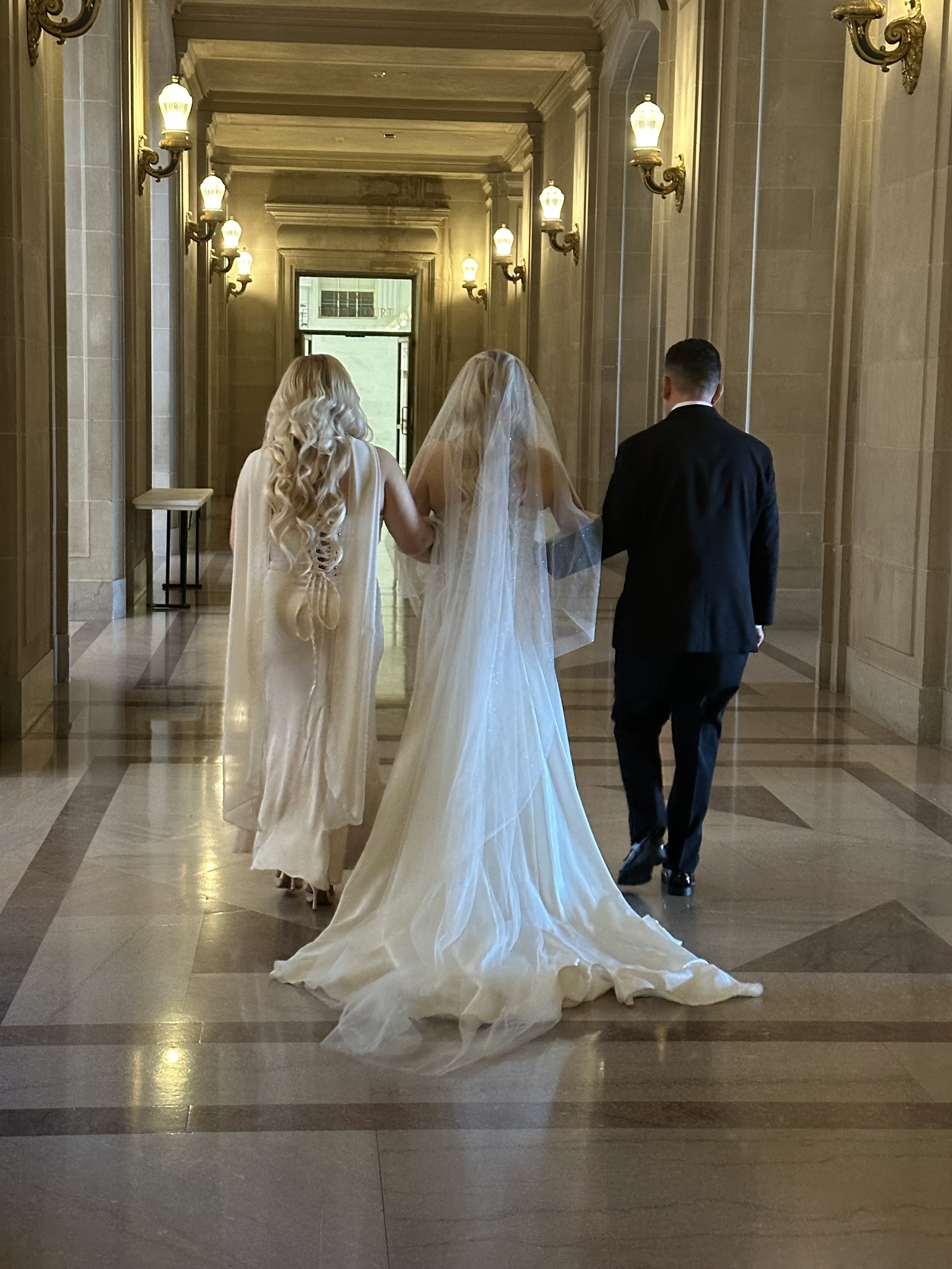 A wedding scene with a bride in a white wedding gown and veil, walking beside a woman with long blonde hair in a cream-colored dress, and a man in a black tuxedo in a grand, well-lit corridor with ornate wall sconces and marble floors.