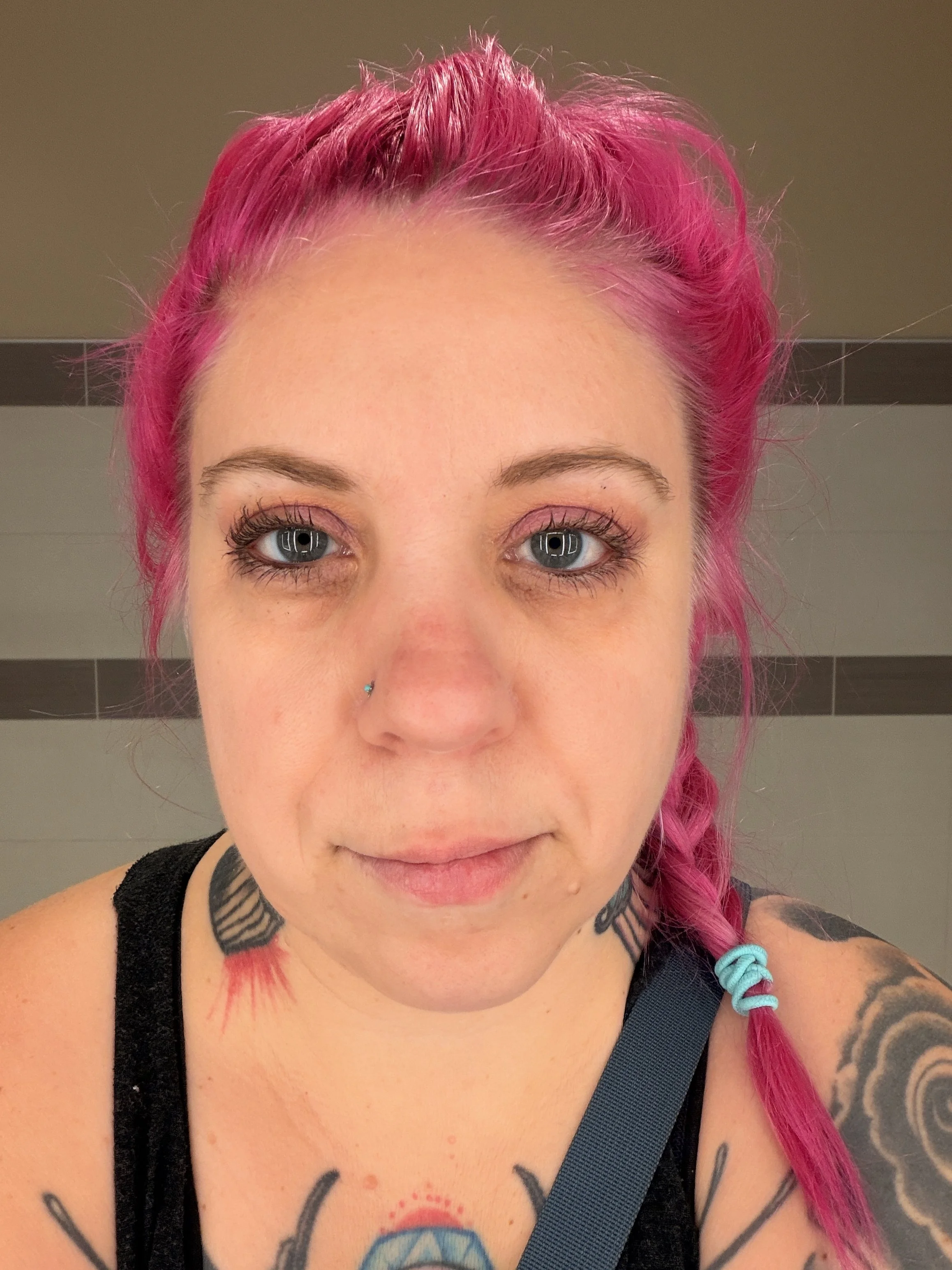 A woman with bright pink hair styled in a braid, wearing a black top, with visible tattoos on her neck and shoulders, and a small nose piercing. She has blue eyes and is looking directly at the camera.