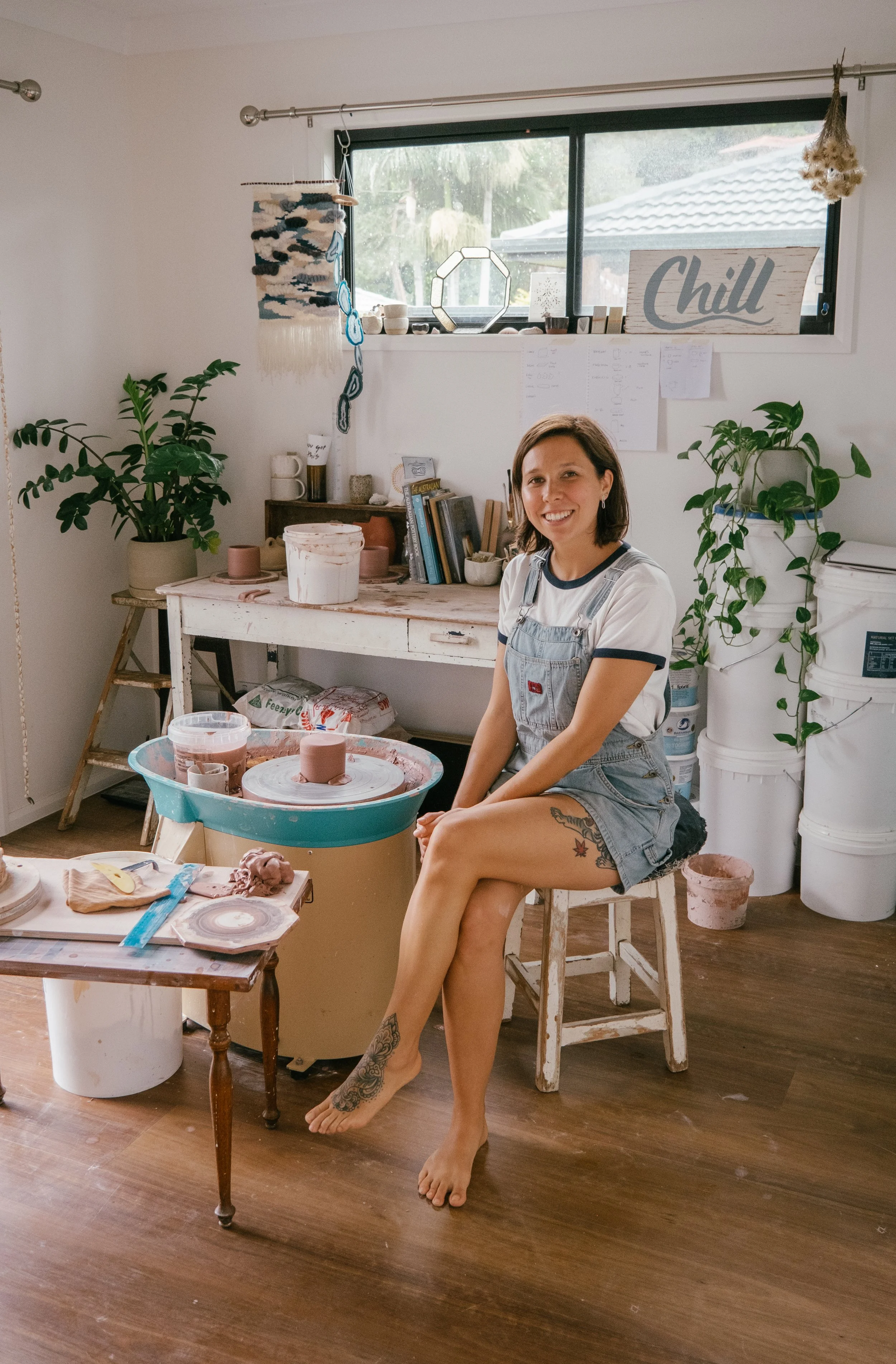 A woman with tattoos on her leg and arm, wearing overalls and a striped t-shirt, sitting barefoot on a stool in an art studio with pottery and plants.