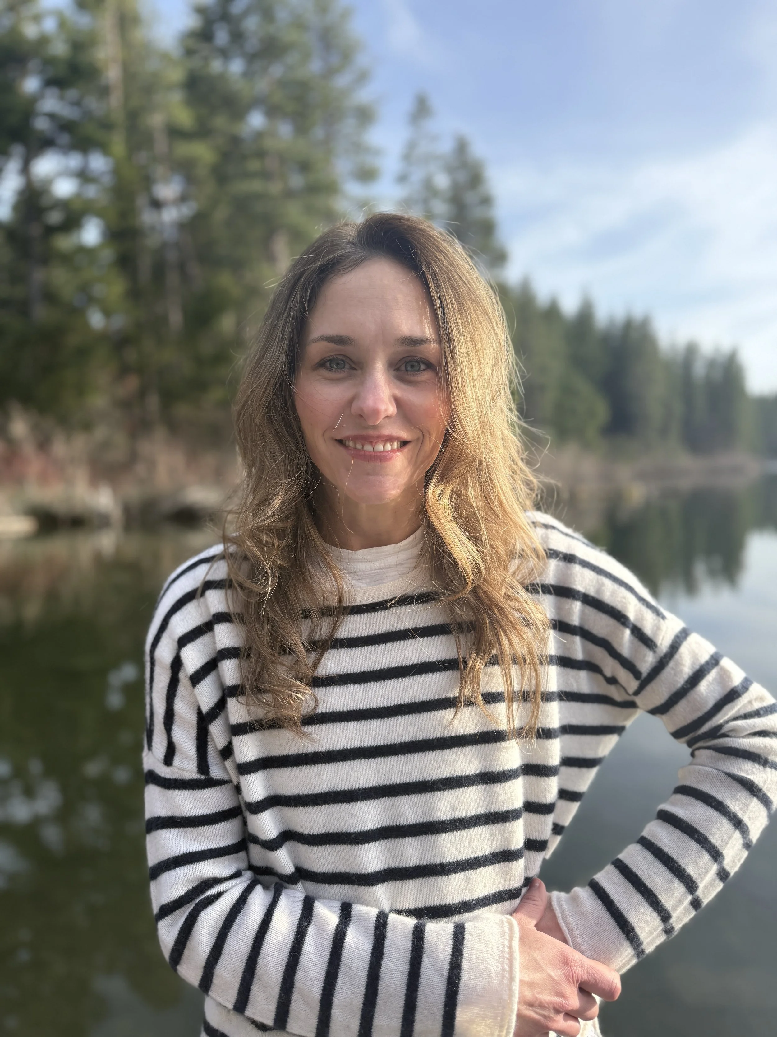 A woman with wavy brown hair and a striped sweater stands outdoors near a body of water with a forest and clear sky in the background.
