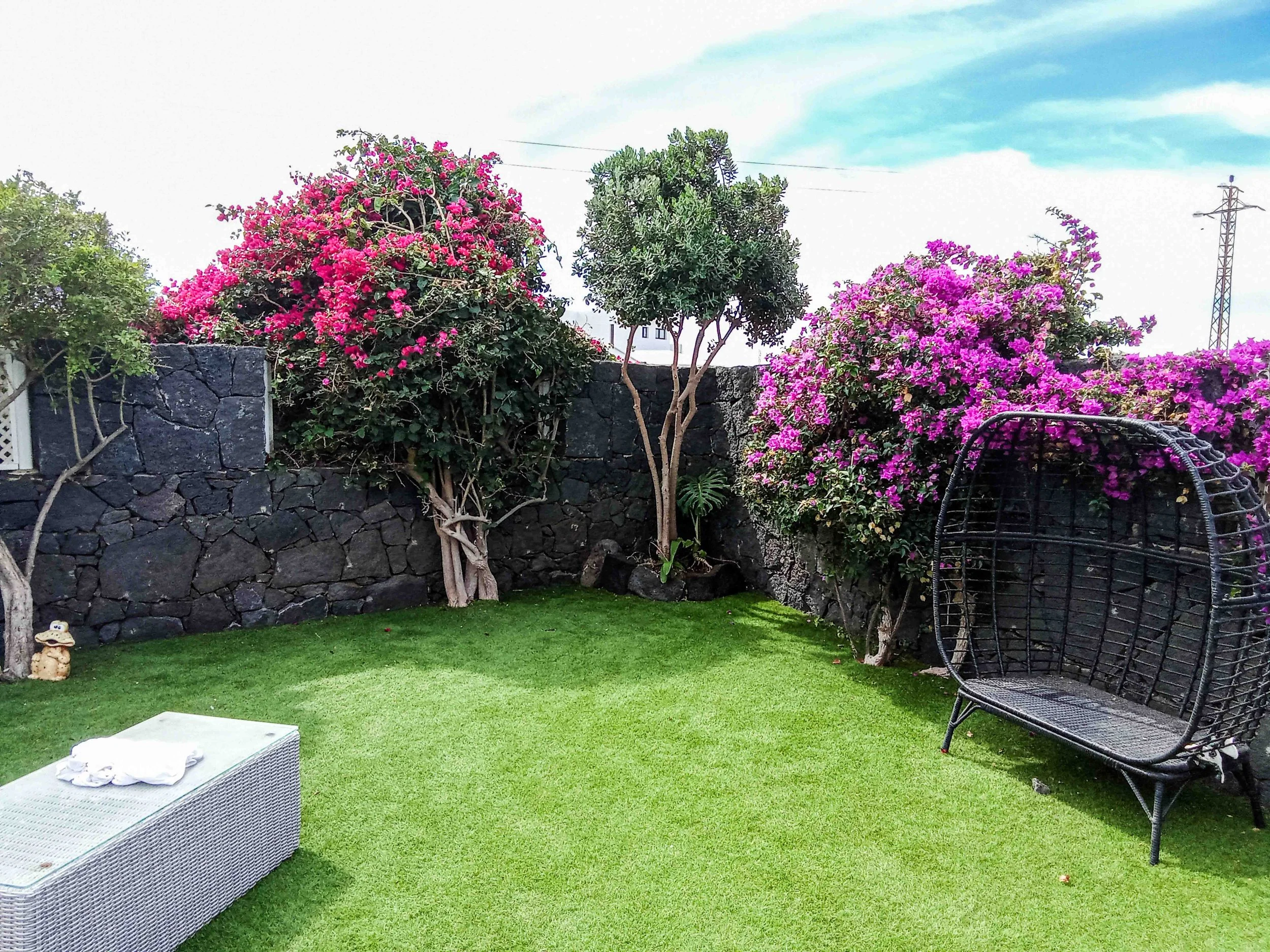 Backyard garden with green artificial grass , flowering pink and purple bushes, a black stone wall, a hanging black wicker chair, a small white wicker table with a towel, a small figurine of a dog, and a partly cloudy sky.