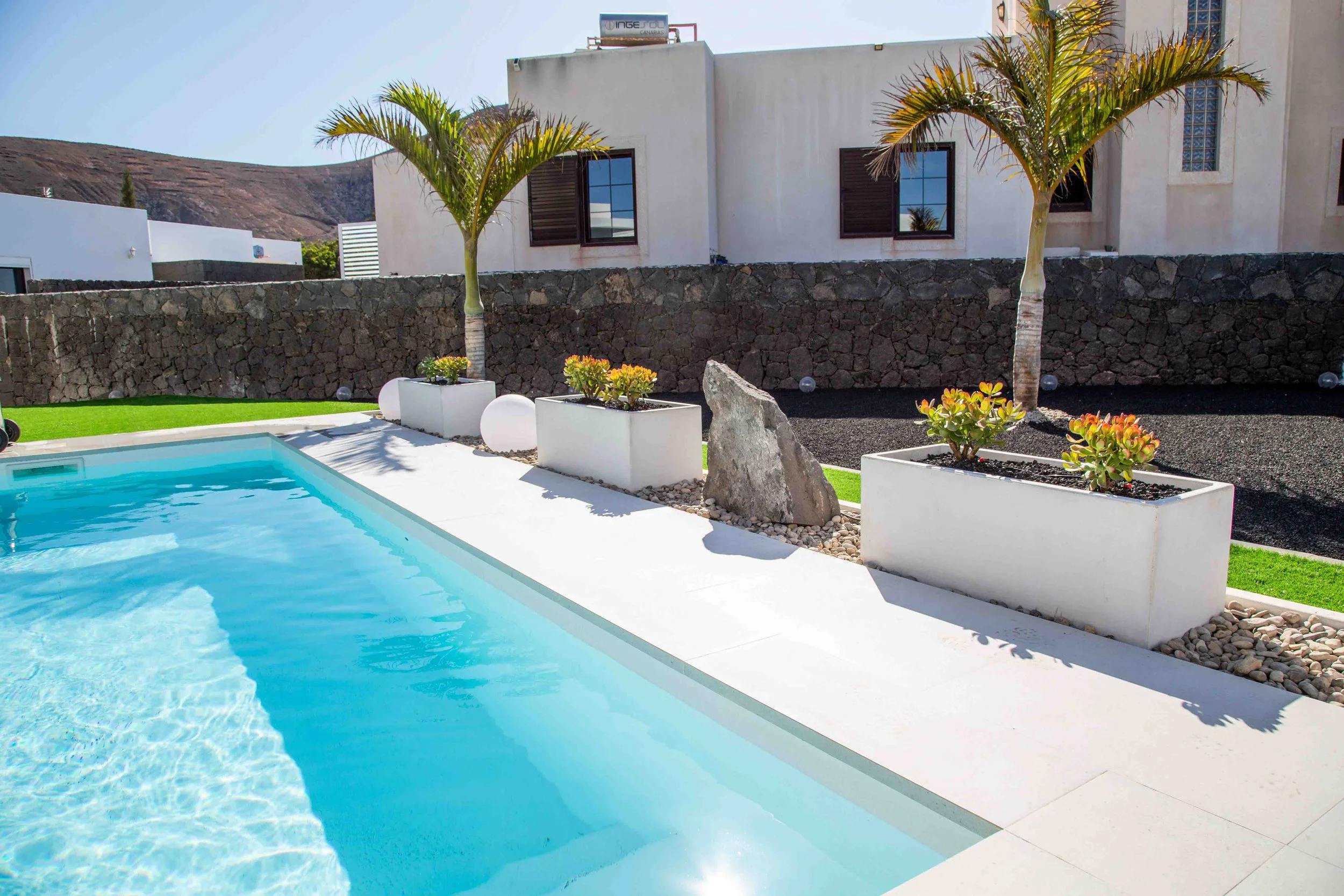 Luxury modern swimming pool with clear blue water, surrounded by white concrete, holiday garden with palm trees, potted plants, rocks, and decorative lighting in front of a contemporary white house with dark brown windows and shutters, and a stone wall.