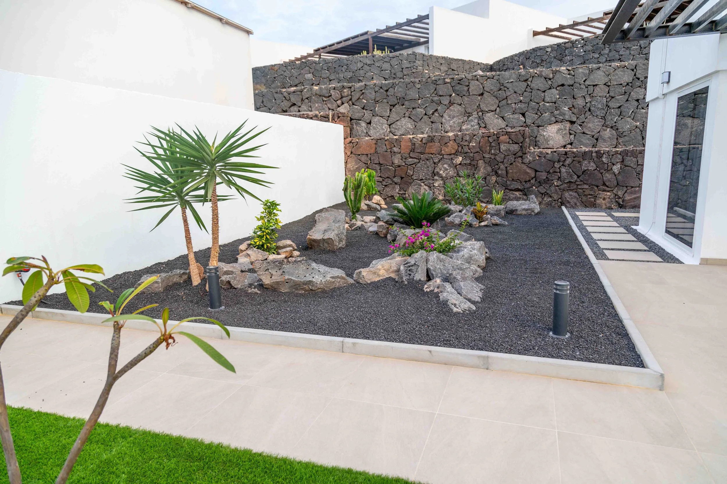 Modern backyard with white walls, black gravel garden bed with large rocks, green plants, and a stone wall background, near a patio area with a door.